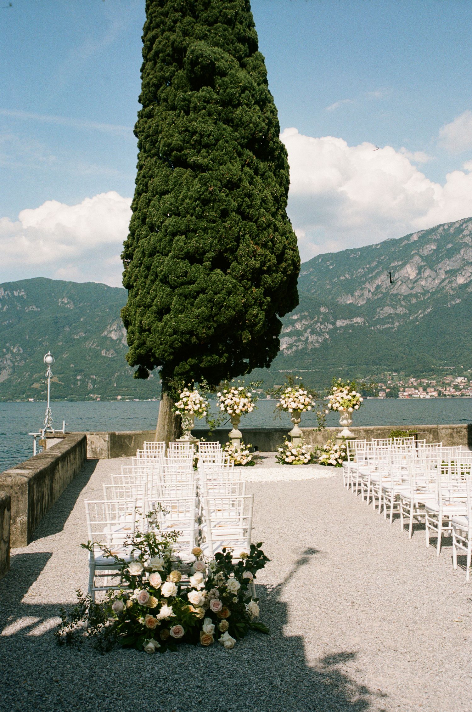 Elegant outdoor wedding ceremony setup on Lake Como terrace with white chairs, floral arrangements, and a tall cypress tree backdrop.