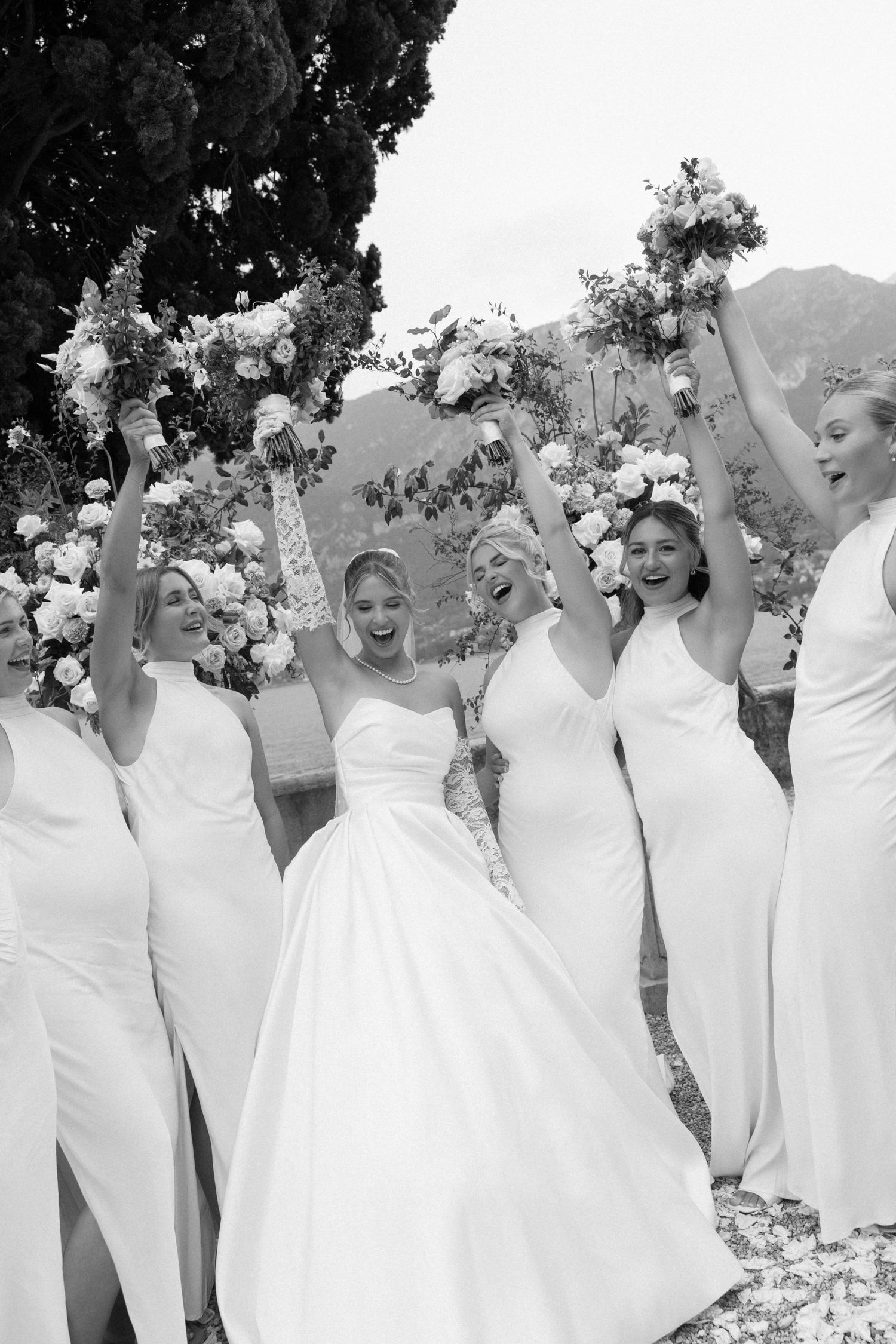 Bride and bridesmaids in white gowns joyfully raising bouquets outdoors at a wedding celebration.
