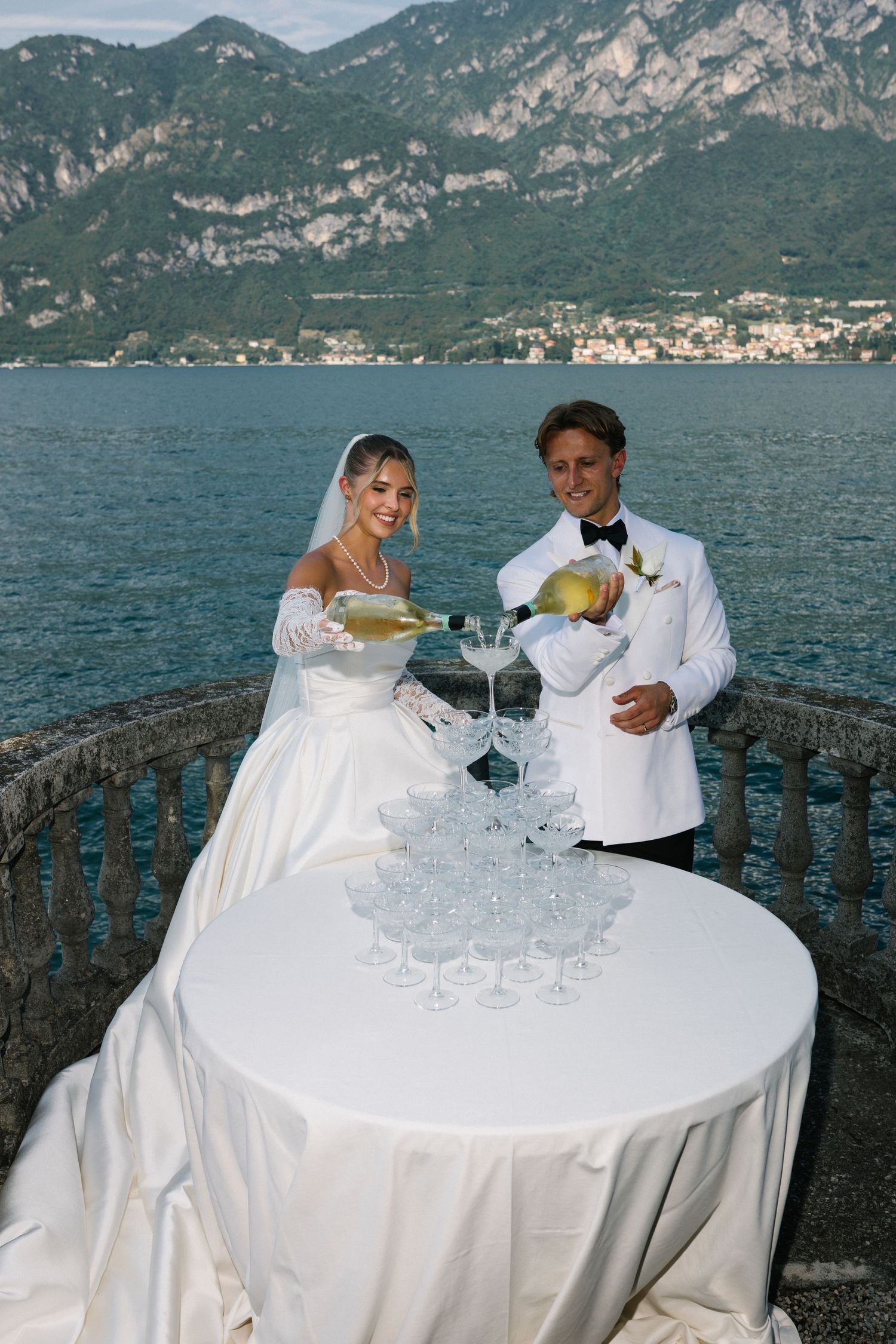 Bride and groom pouring champagne tower on lakeside terrace with mountain backdrop at elegant Italian wedding.