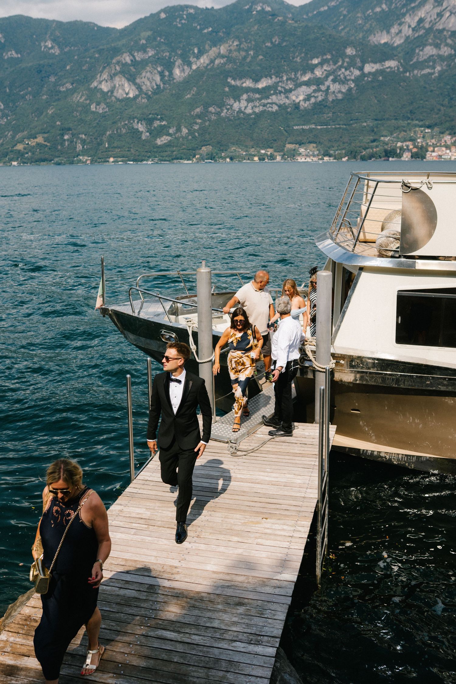 Wedding guests disembark from a luxury yacht onto a wooden dock with stunning mountain lake views.