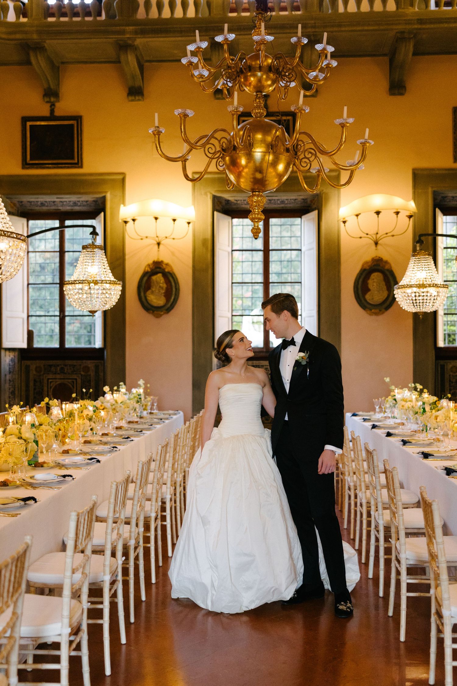 Bride and groom stand together in an ornate Italian villa reception hall with gold chandeliers and long floral-adorned tables.