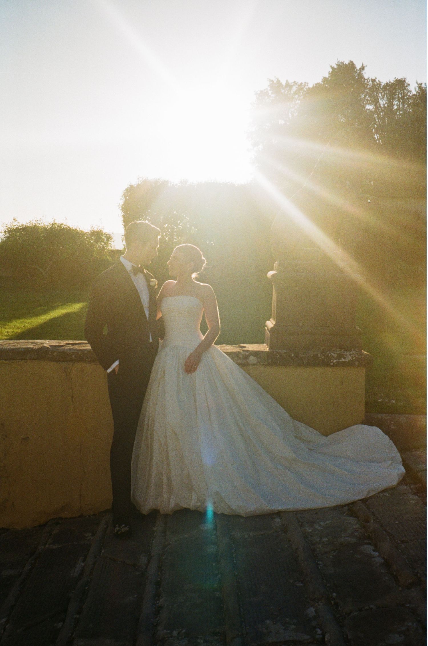 A bride and groom share a romantic moment backlit by golden sunset rays at an outdoor wedding venue.