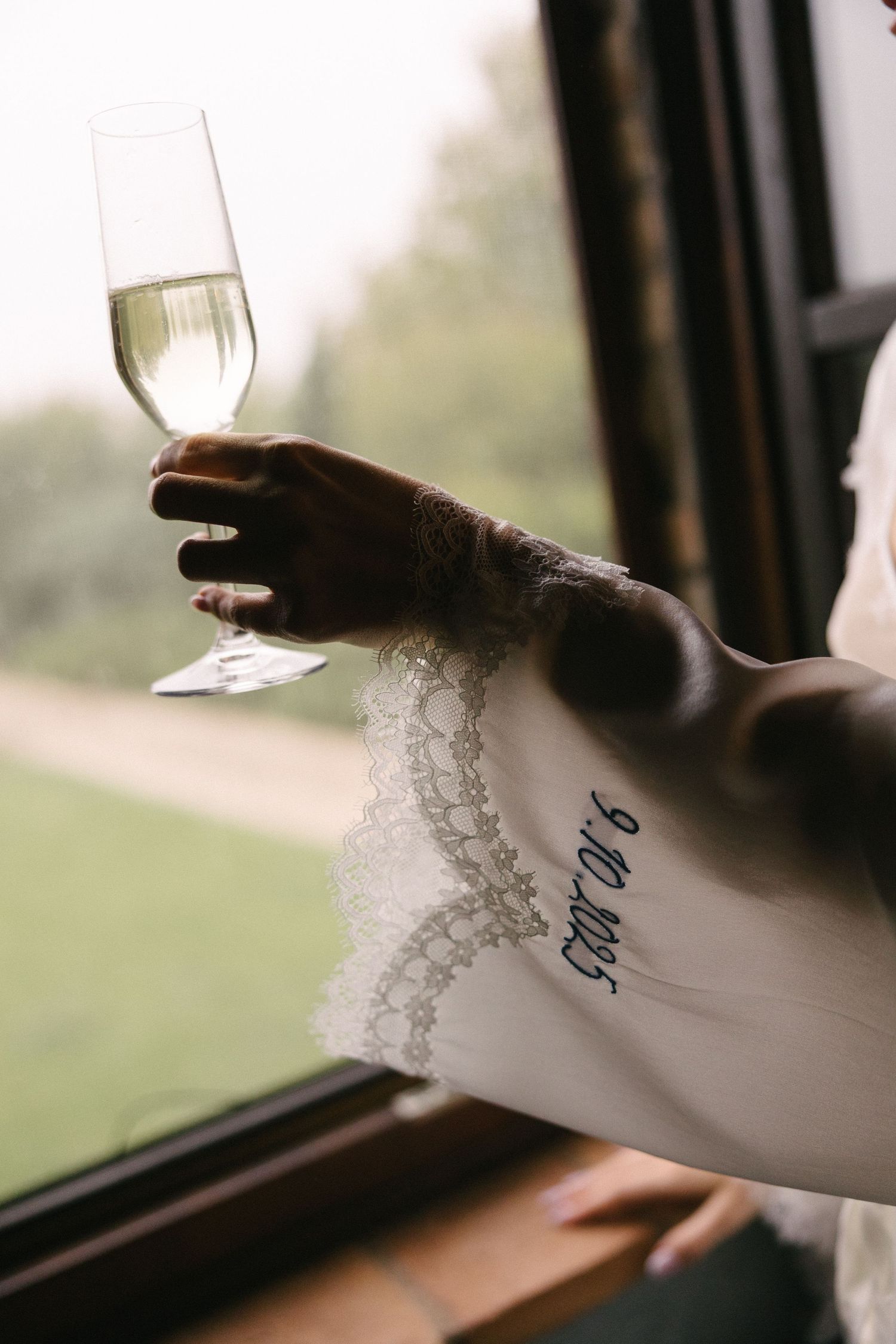 Bride holding champagne flute with lace wedding dress sleeve embroidered with wedding date 9.10.2025 near window.