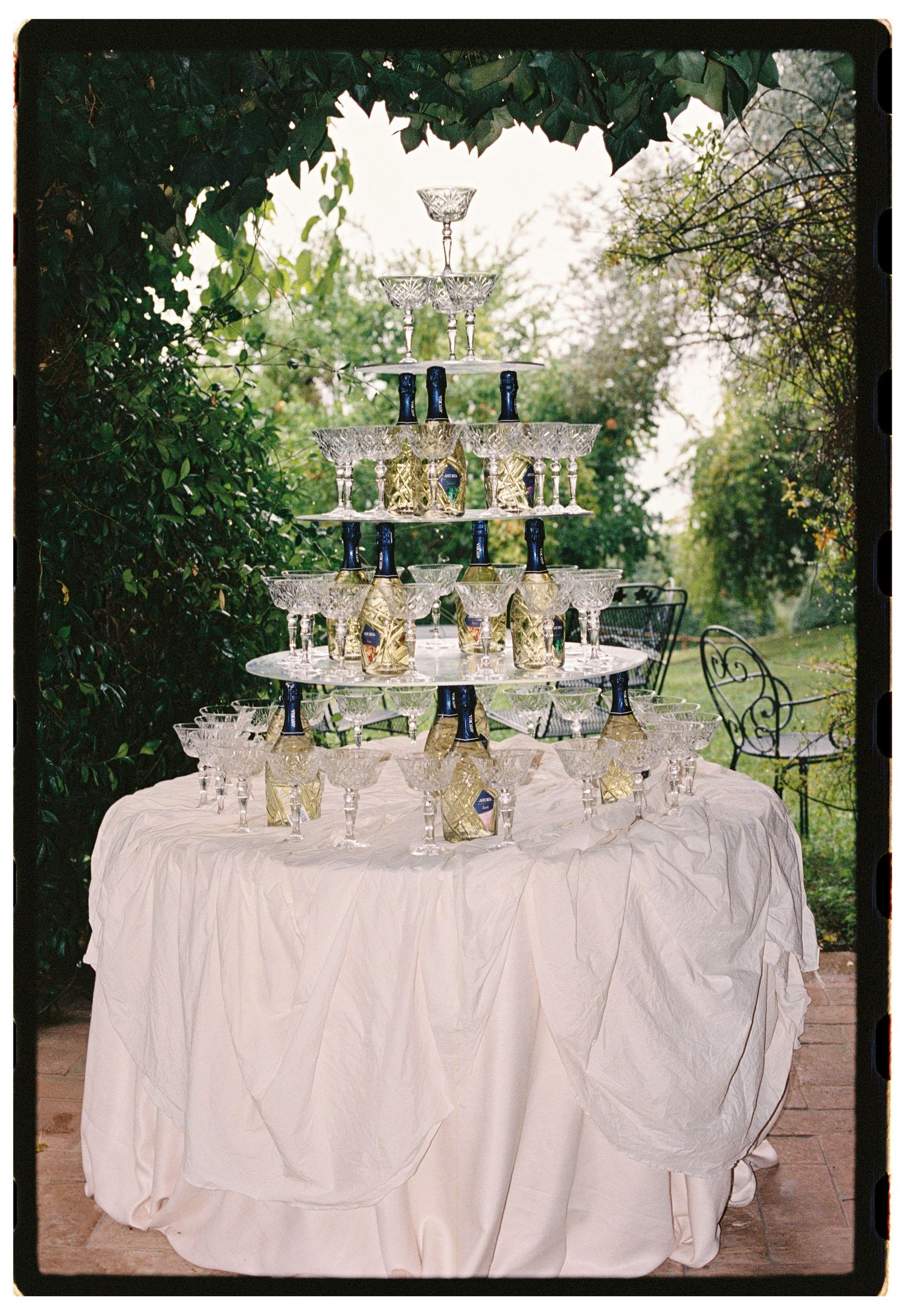 Elegant champagne tower with crystal glasses and blue bottles on a white draped table in a lush garden setting.