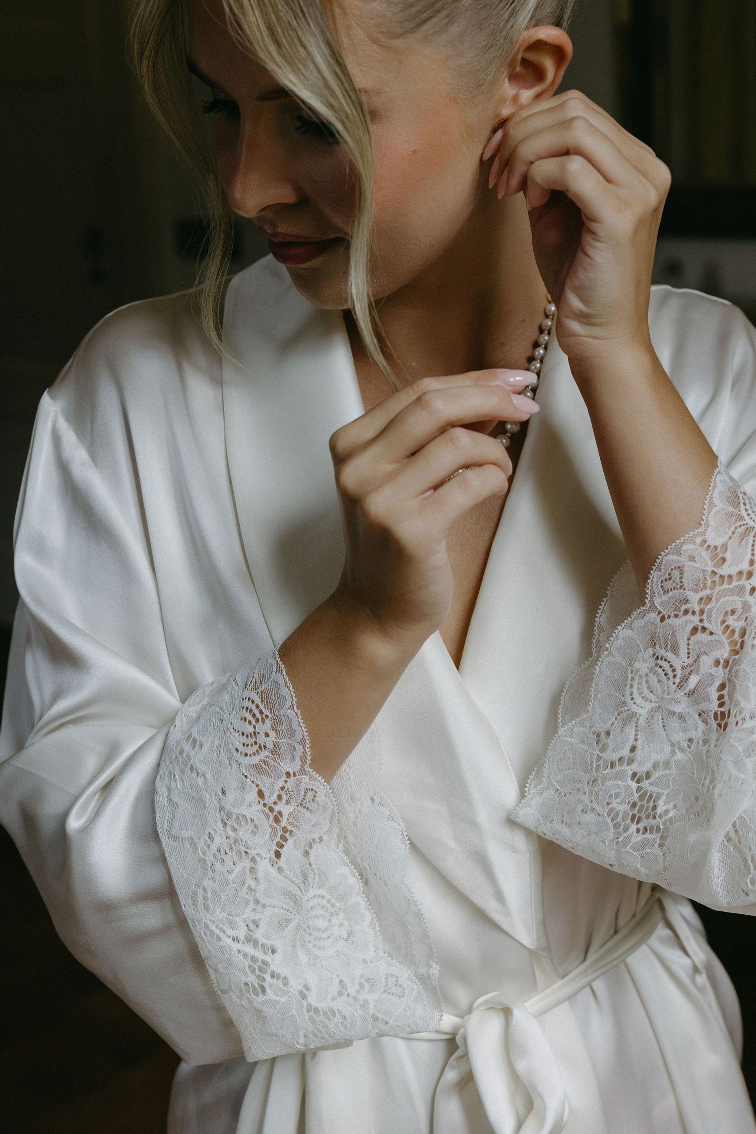 Blonde bride in white satin lace robe putting on pearl earrings while getting ready for wedding day.