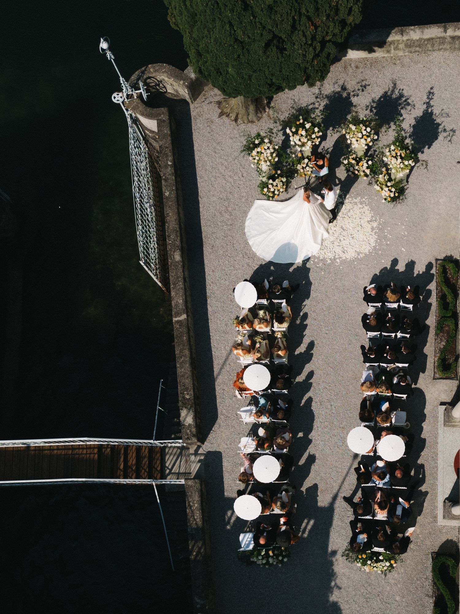 Aerial drone view of an outdoor wedding ceremony with guests seated in white chairs near a waterfront venue.