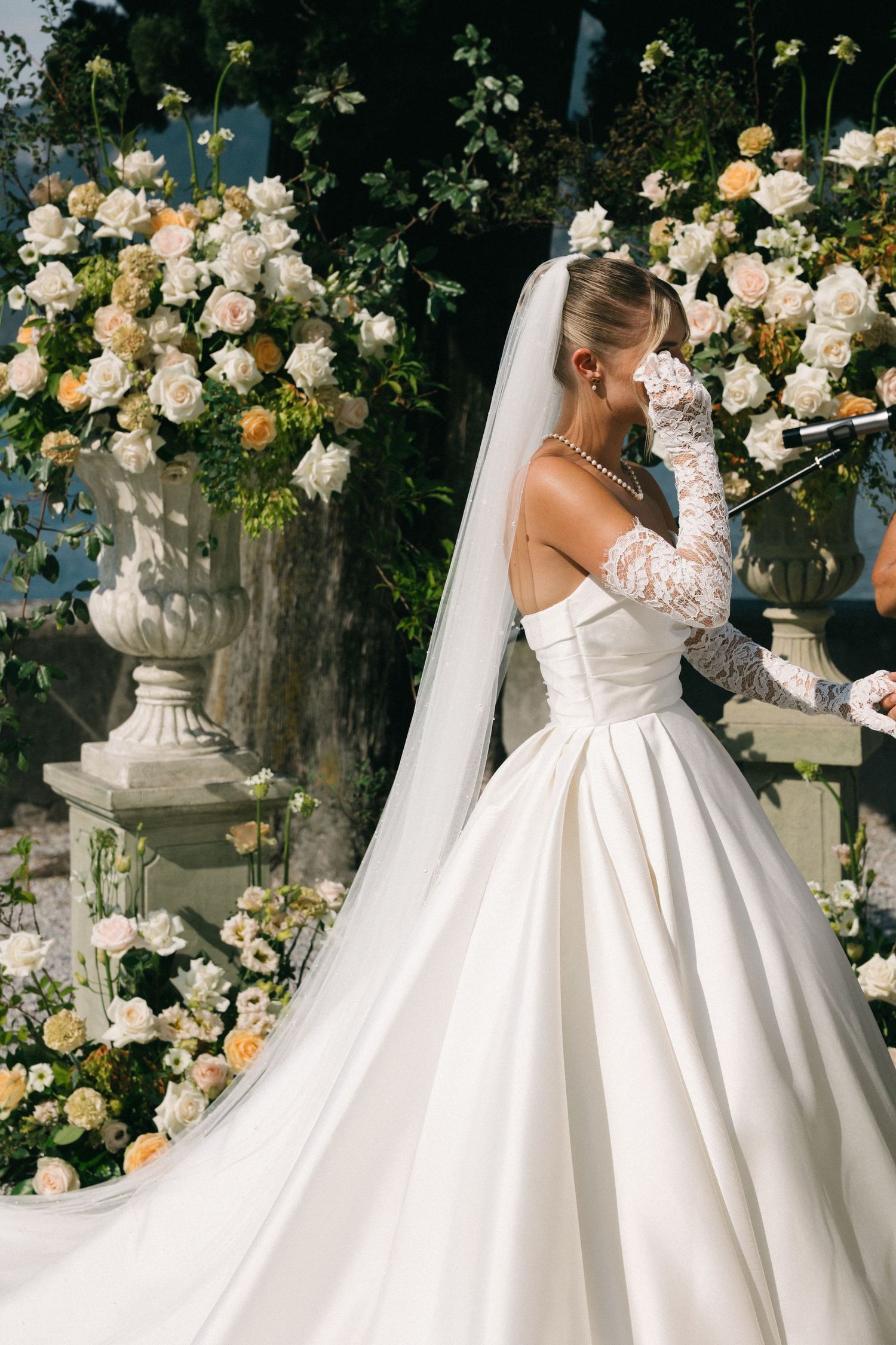 Bride in elegant off-shoulder ballgown with lace gloves and veil stands among lush white and peach floral arrangements.