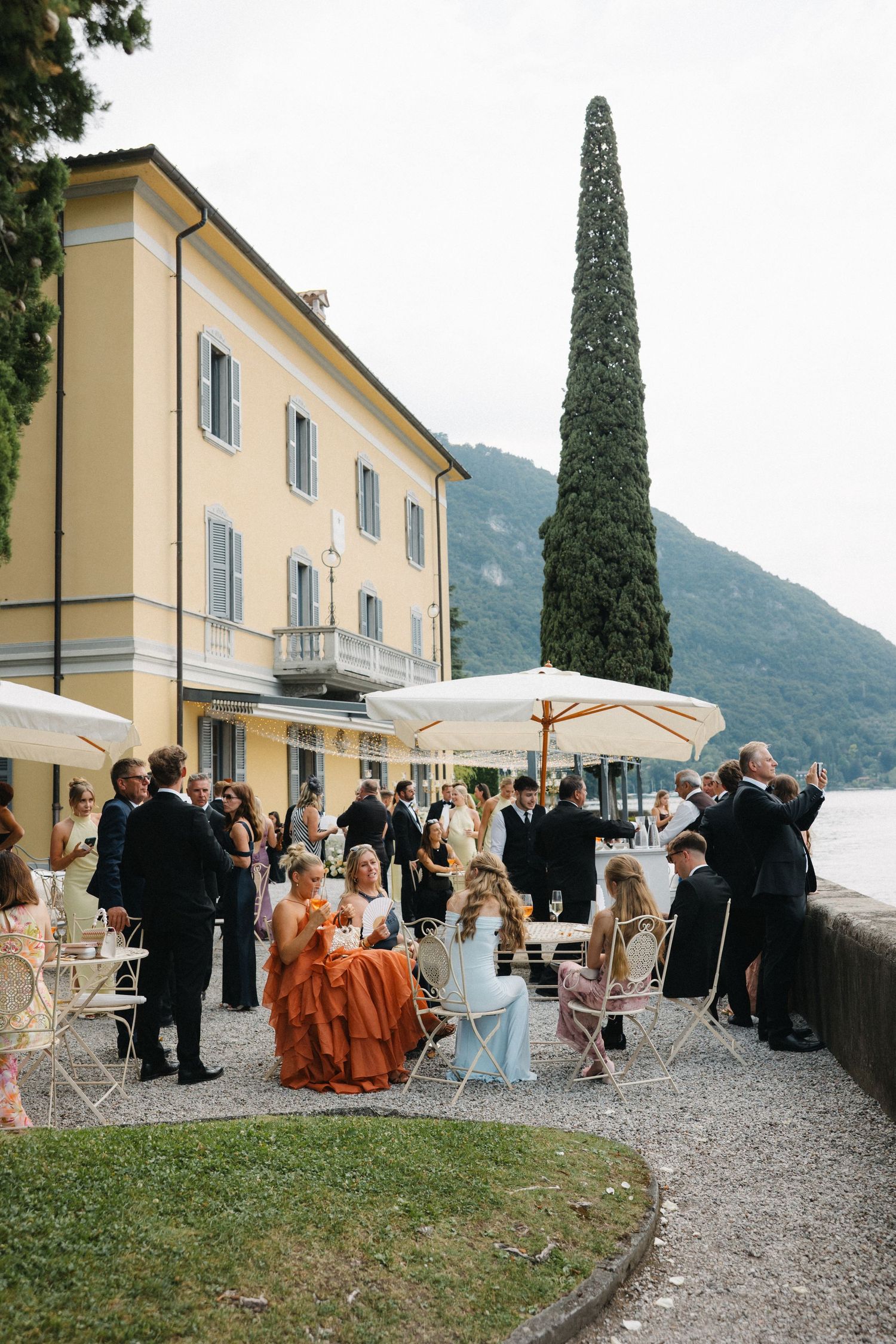 Elegant outdoor wedding reception at Italian lakeside villa with guests in formal attire seated at white tables near Lake Como.