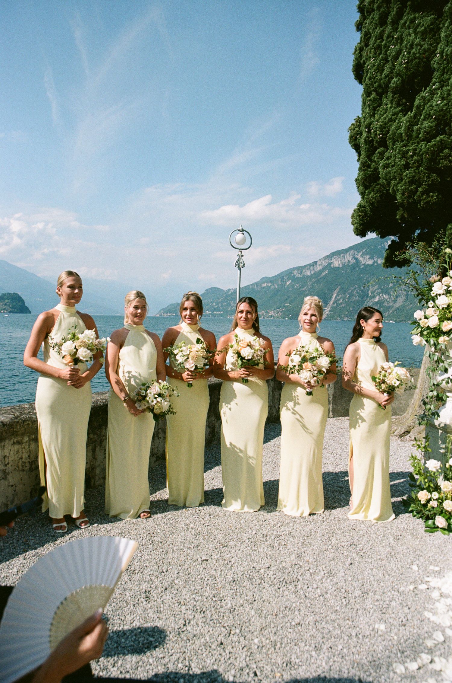 Six bridesmaids in yellow halter gowns holding bouquets pose by a lakeside terrace with mountain views.