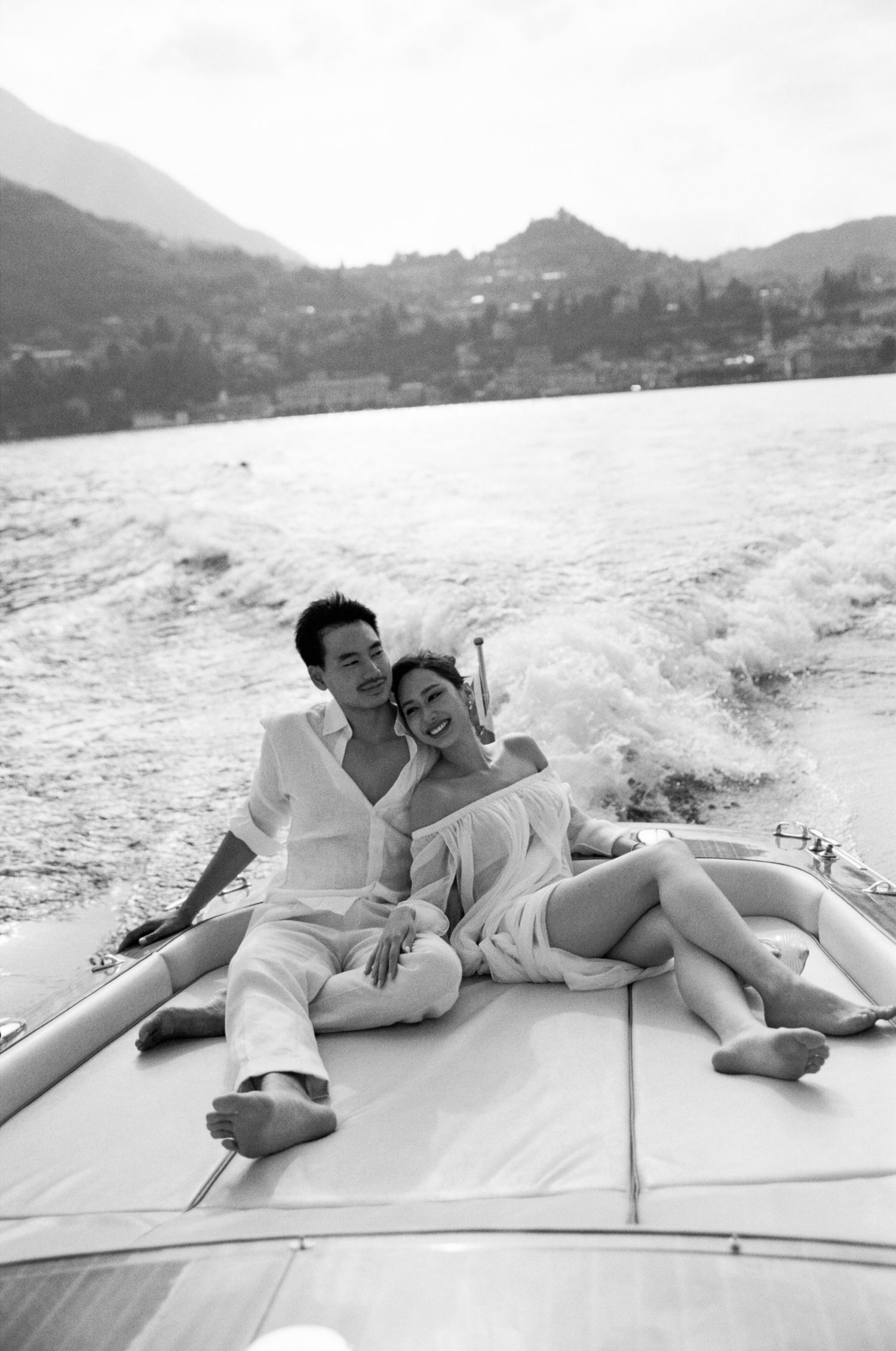 Newlyweds relax together on boat deck laughing with scenic mountain lake view in black and white photo.