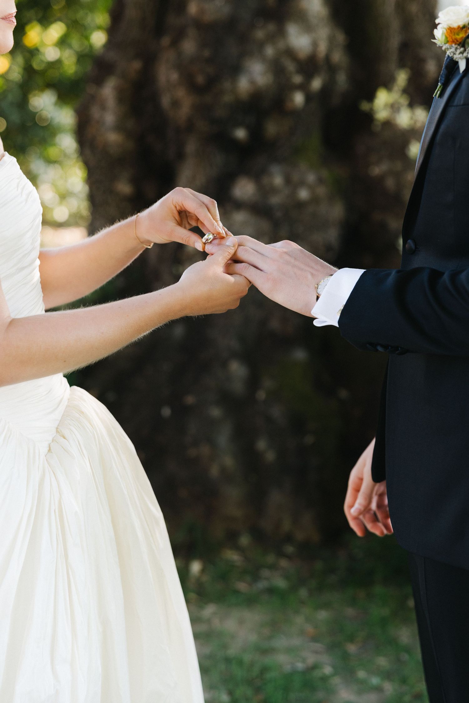 Bride placing a wedding ring on grooms finger during an outdoor ceremony surrounded by trees.