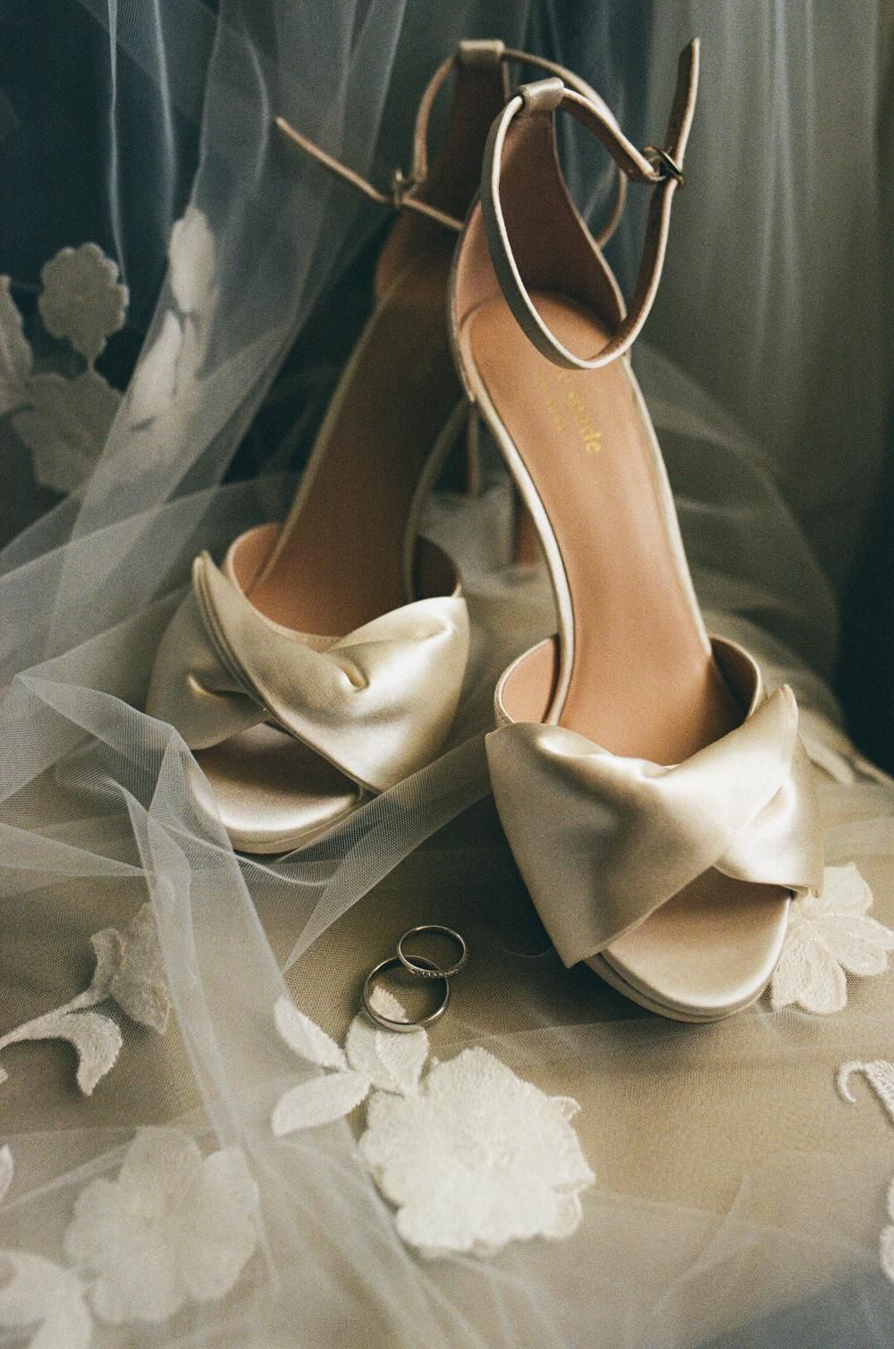 Elegant champagne satin bridal heels with bow detail and ankle strap beside wedding rings on lace veil.