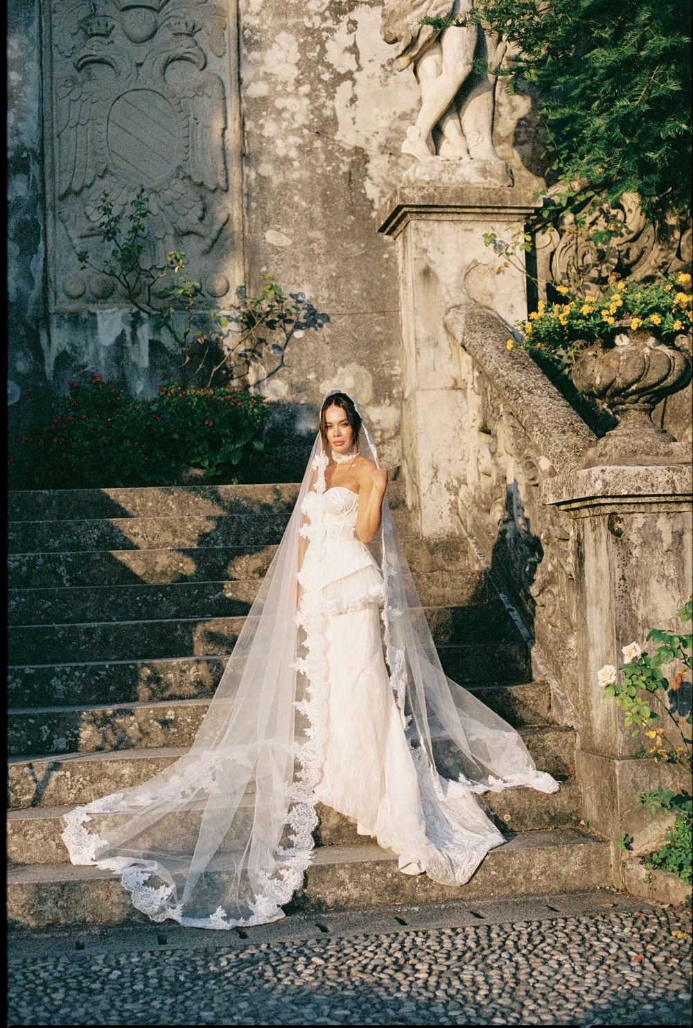 Elegant bride in strapless gown with long lace veil posing on stone steps at historic venue.