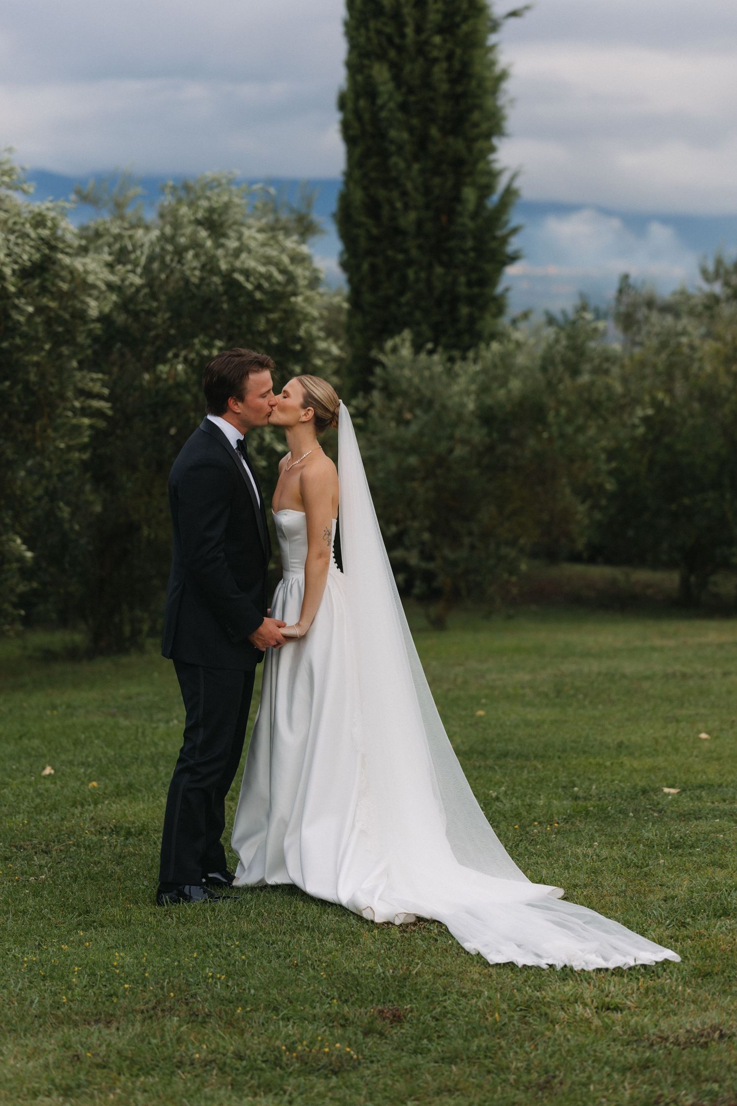 Bride and groom share a kiss on green lawn with cypress trees and olive grove backdrop on their wedding day.
