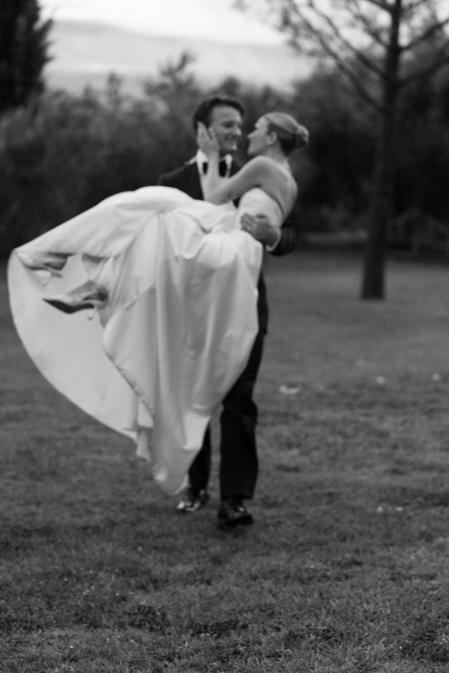 Groom lifts bride in white wedding dress outdoors on grass, black and white romantic wedding photo.