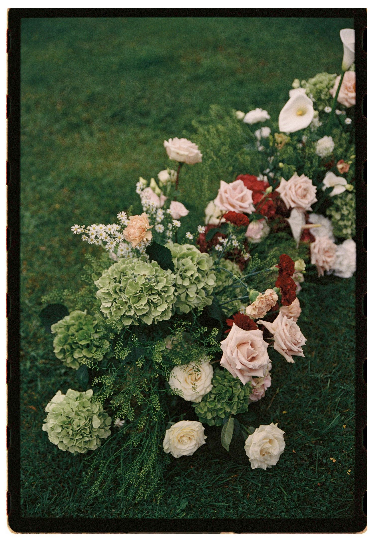 Lush floral arrangement on green grass featuring pink roses, white hydrangeas, red blooms, and greenery for a garden wedding.
