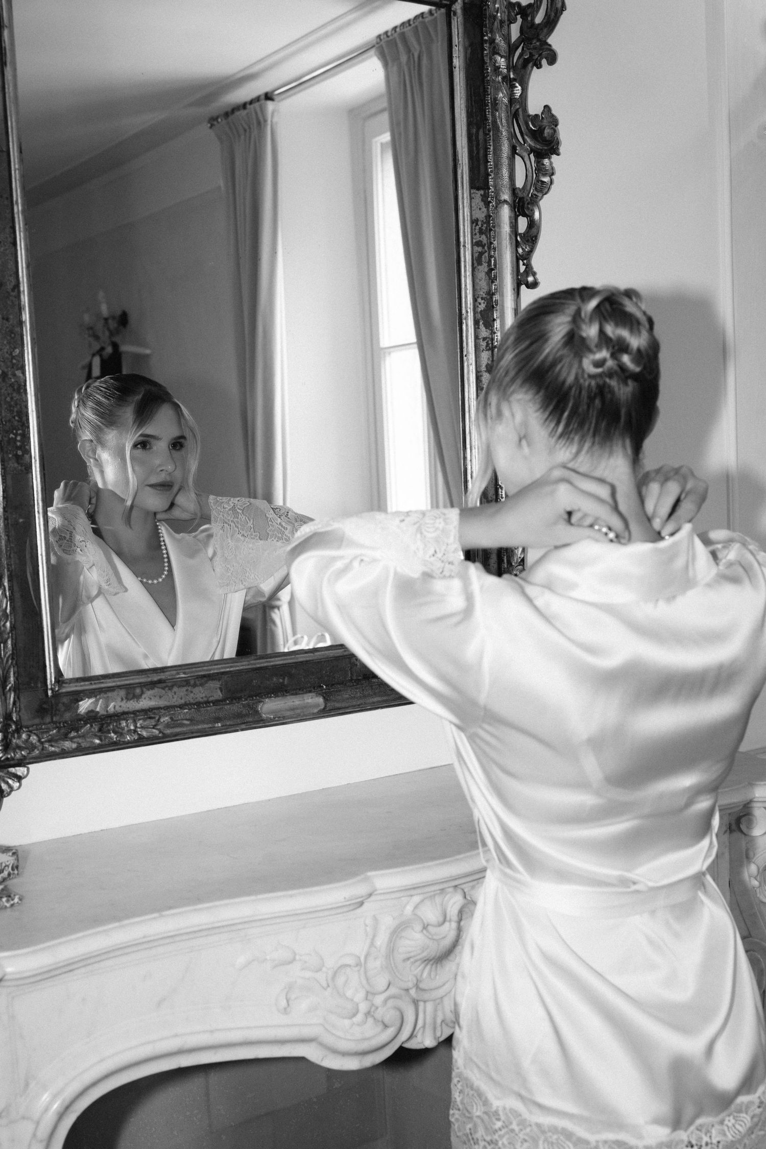 Black and white photo of a bride in a satin robe adjusting her necklace while looking in an ornate mirror.