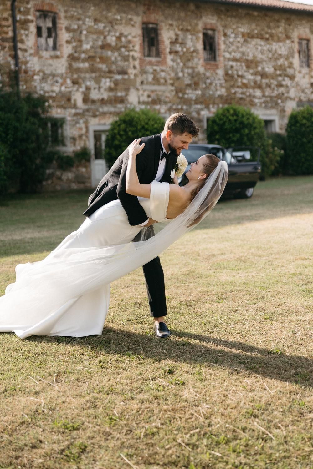 Groom dips bride in white wedding dress and veil outside rustic stone building on sunny day.
