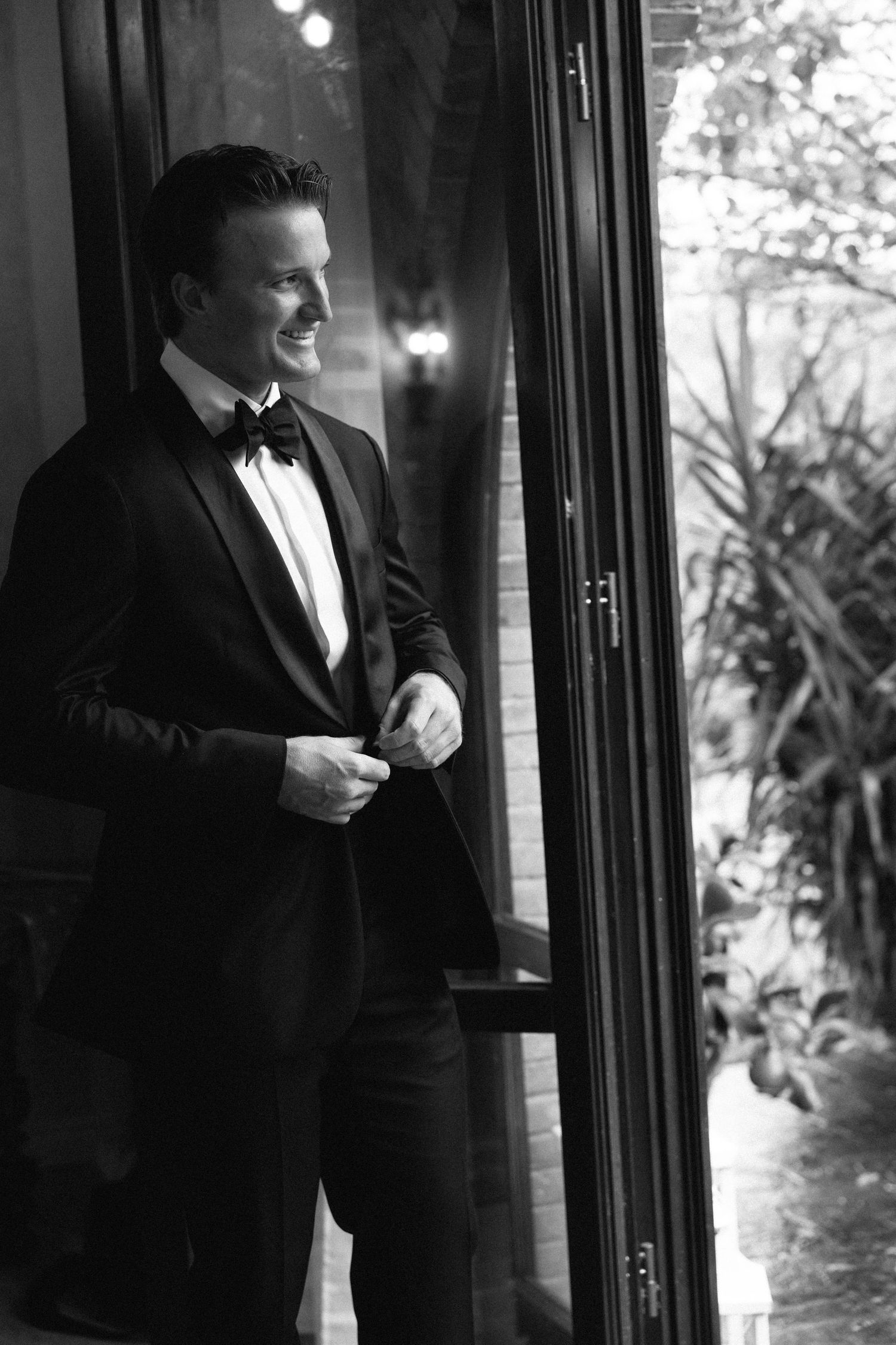Black and white portrait of smiling groom in tuxedo and bow tie standing by glass door.