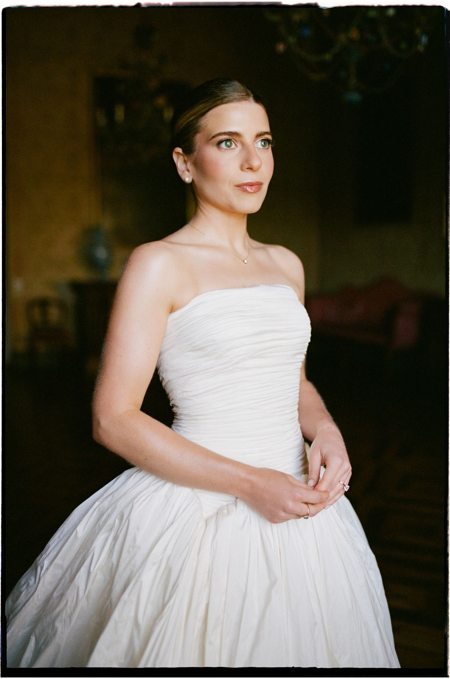 Bride in strapless white ruched ball gown wedding dress poses elegantly in a dimly lit room.