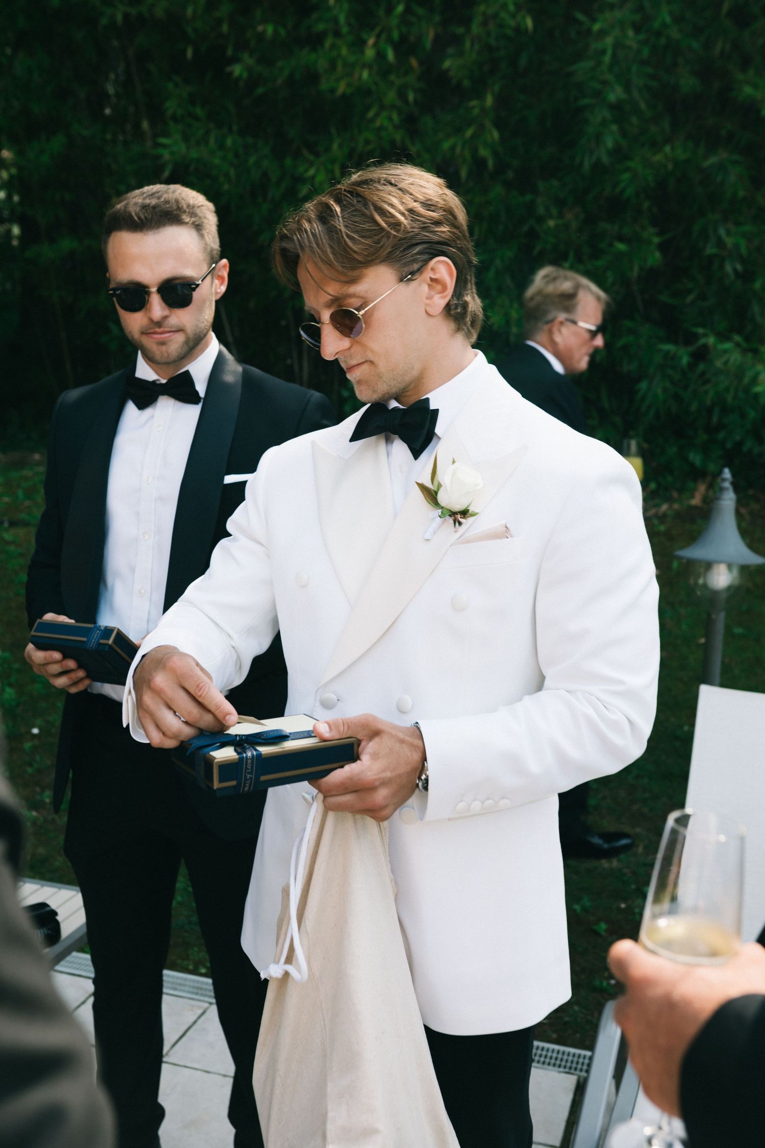 Groom in white tuxedo jacket pours champagne at outdoor wedding reception with groomsmen in black tuxedos.
