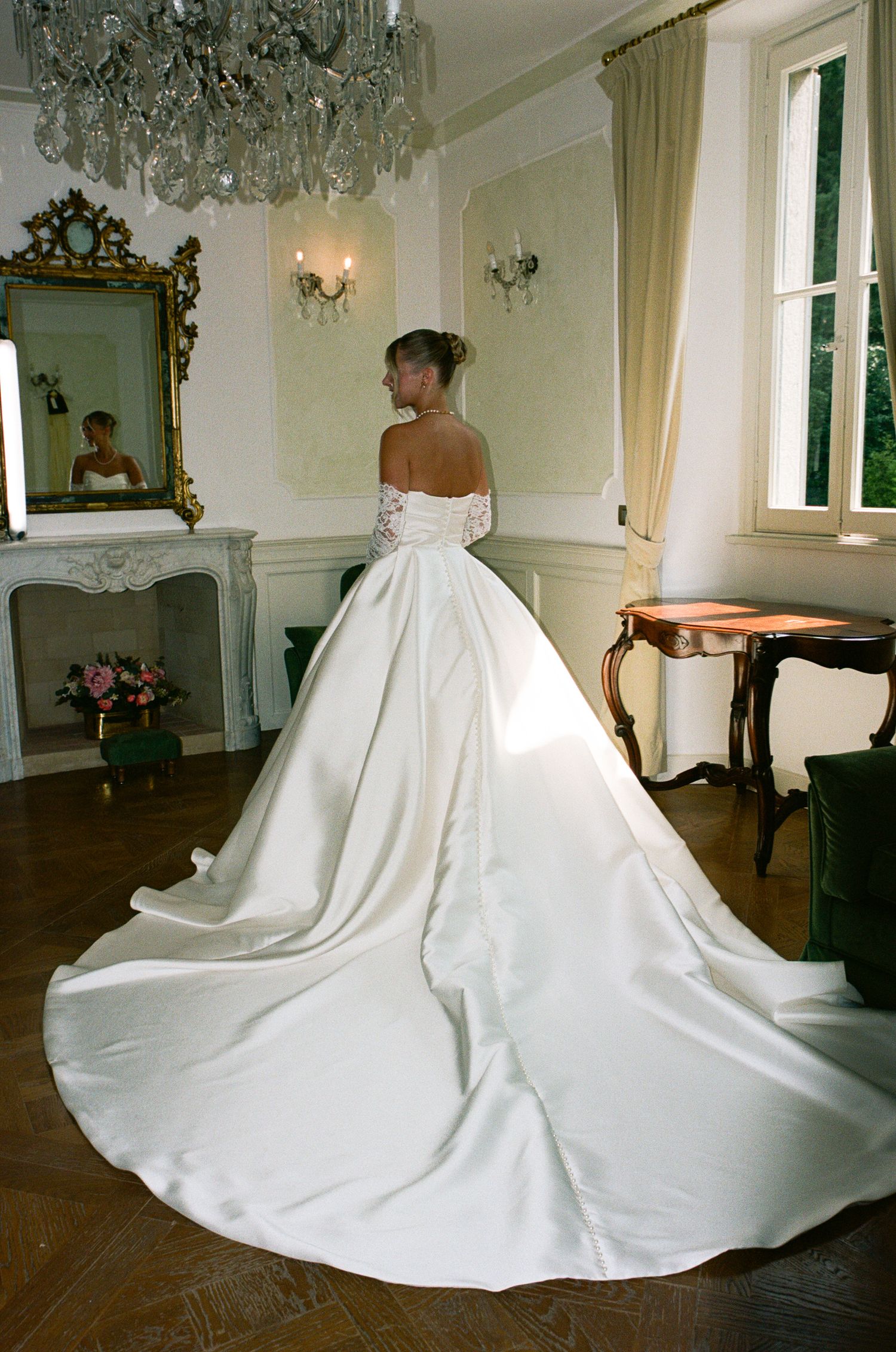 Bride in elegant off-shoulder satin ball gown with long train poses in ornate room with chandelier and fireplace.