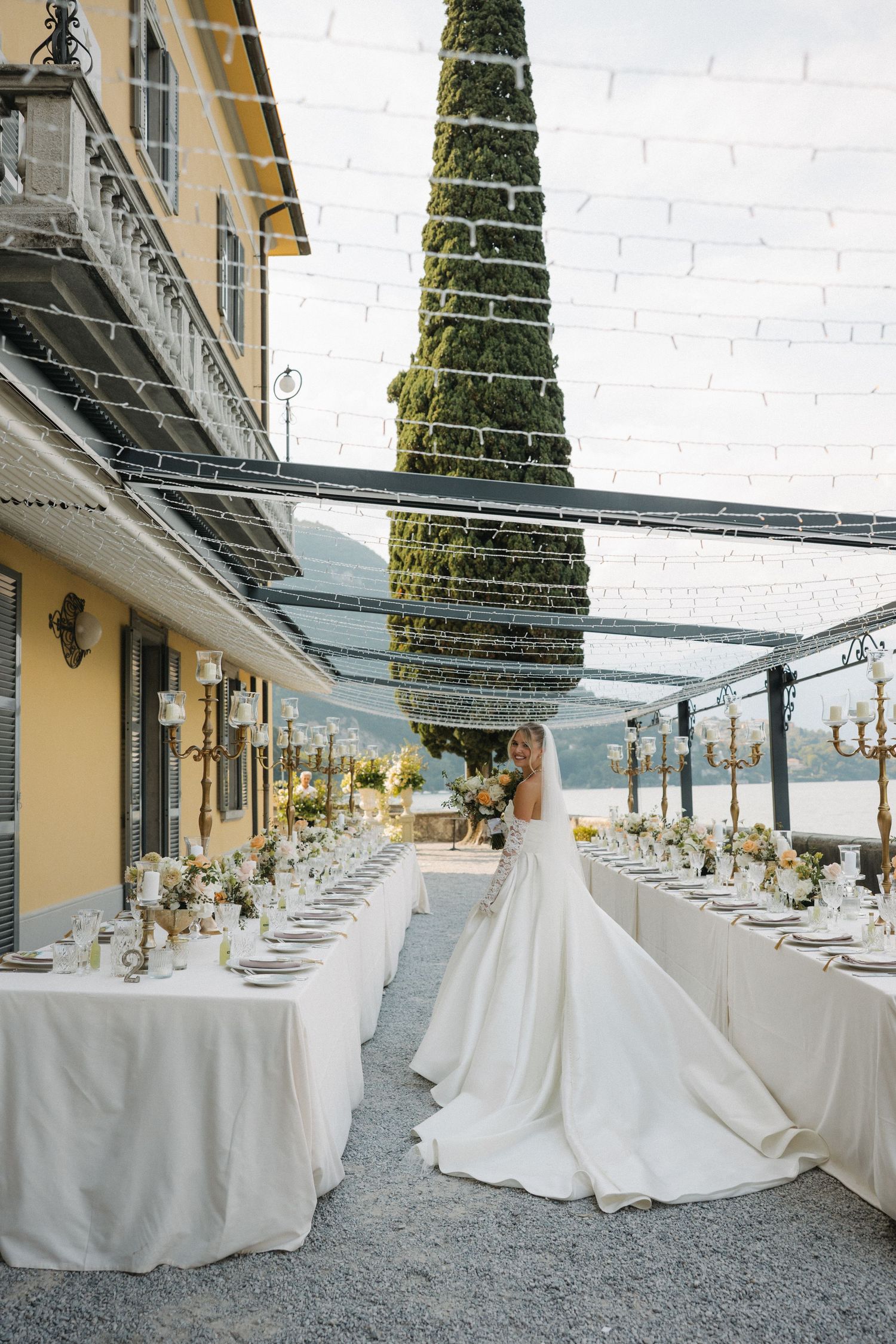 Bride in elegant white gown stands between long reception tables at an outdoor lakeside wedding venue with string lights.