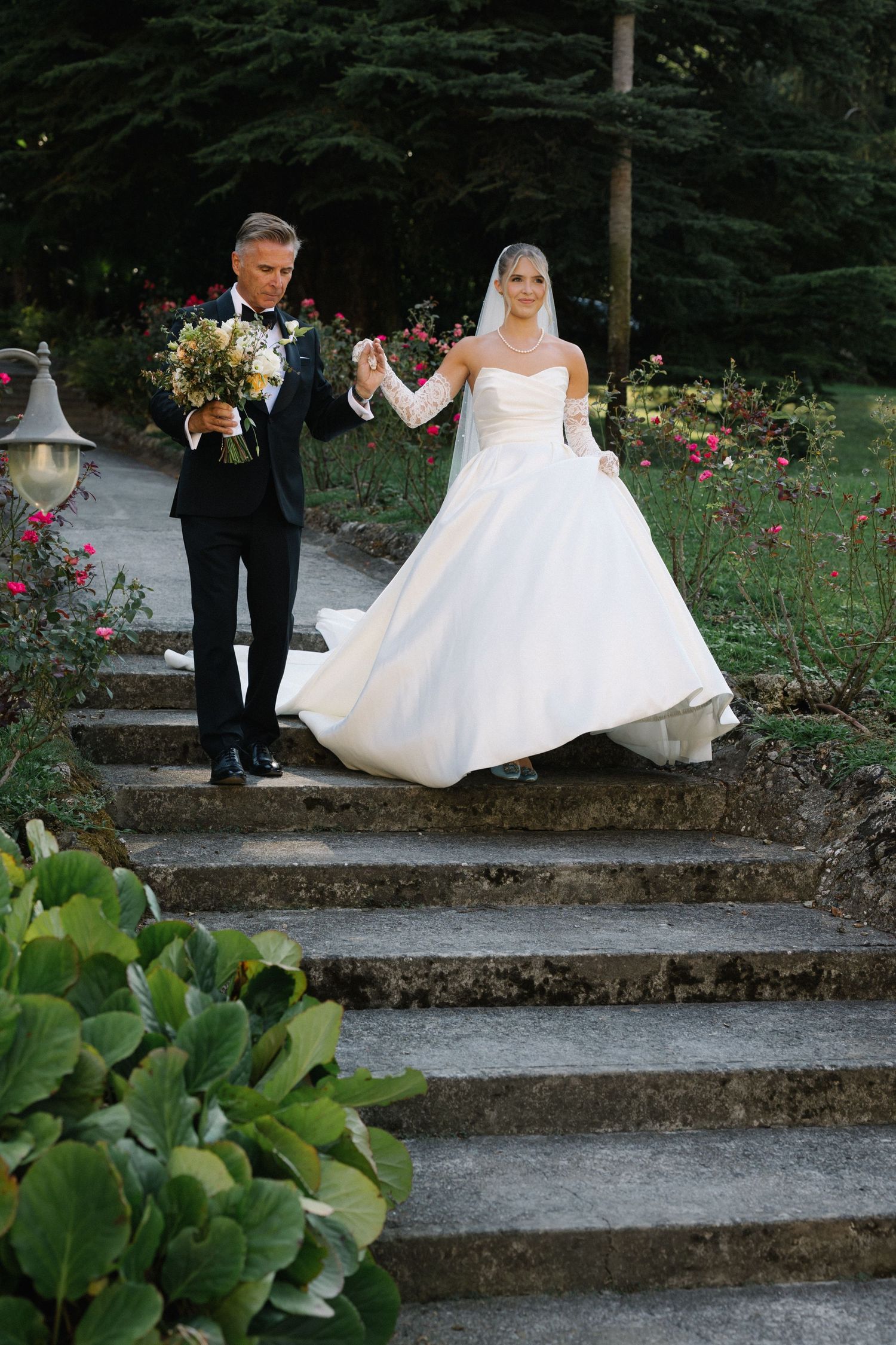 Bride and groom walking down stone steps in a lush garden, bride in white ballgown with veil, groom in black tuxedo.