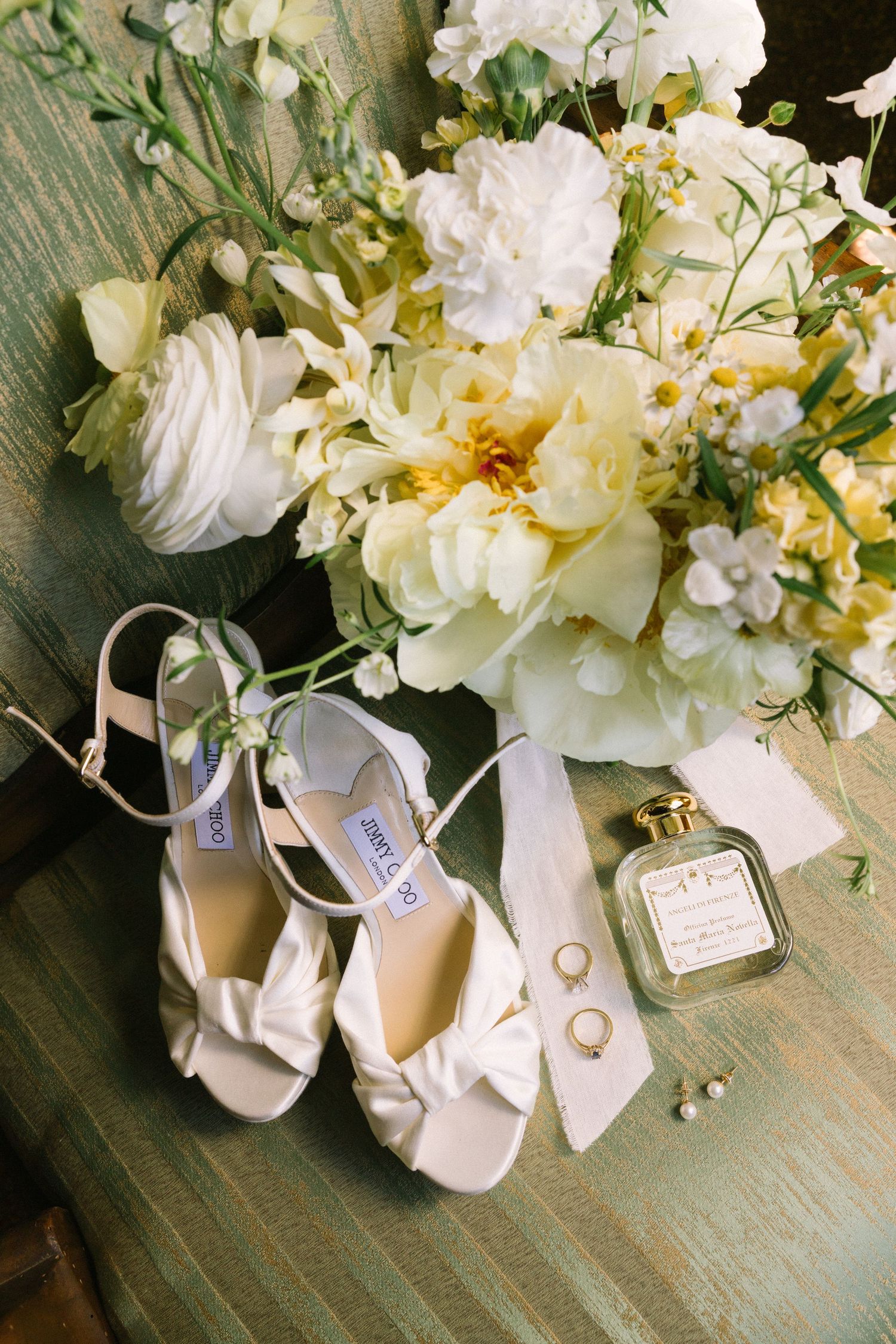 Jimmy Choo white bridal heels displayed with yellow and white floral bouquet, perfume, rings, and pearl earrings.
