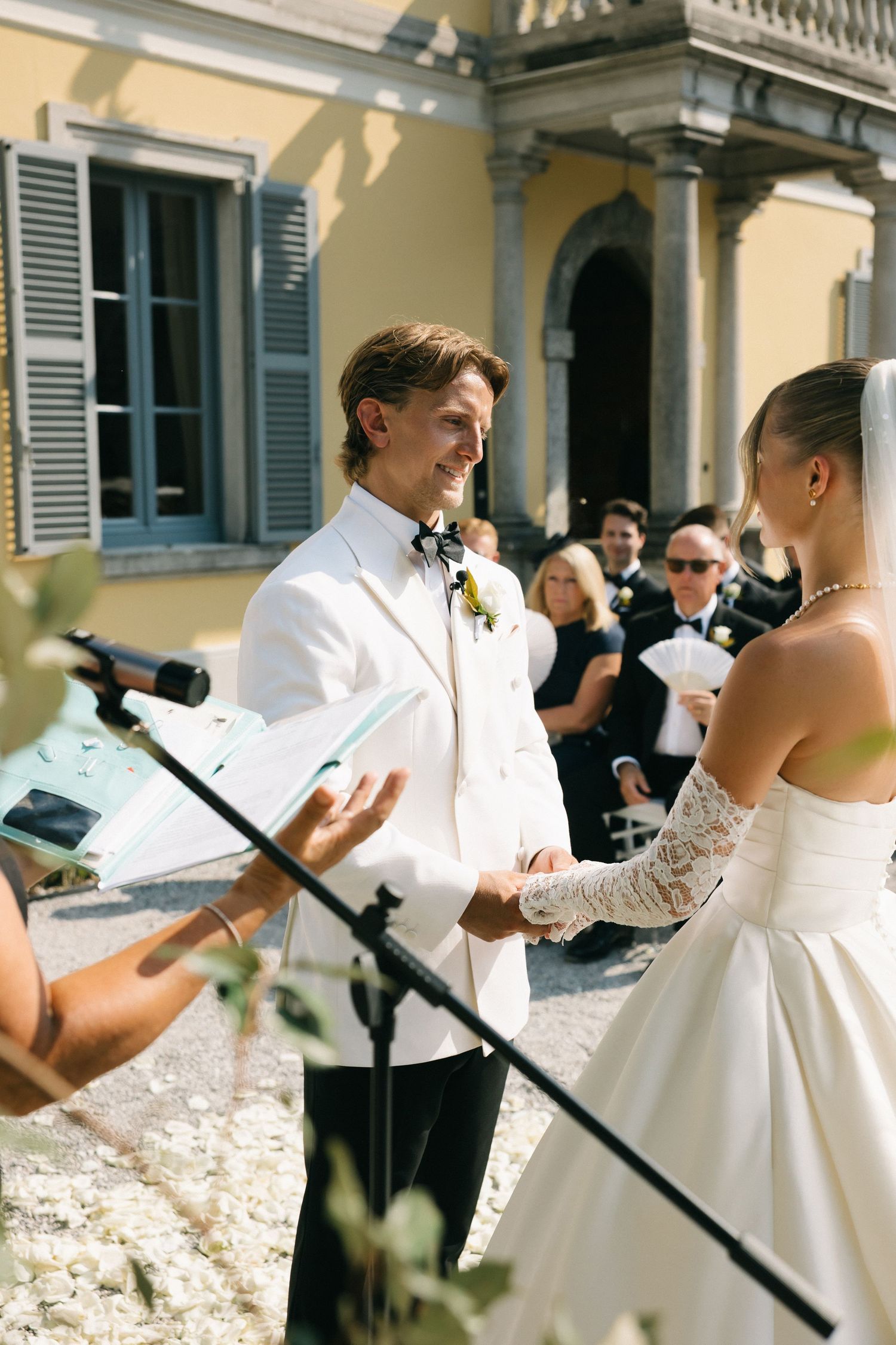 Groom in white tuxedo exchanges vows with bride outdoors at elegant villa wedding ceremony with seated guests.