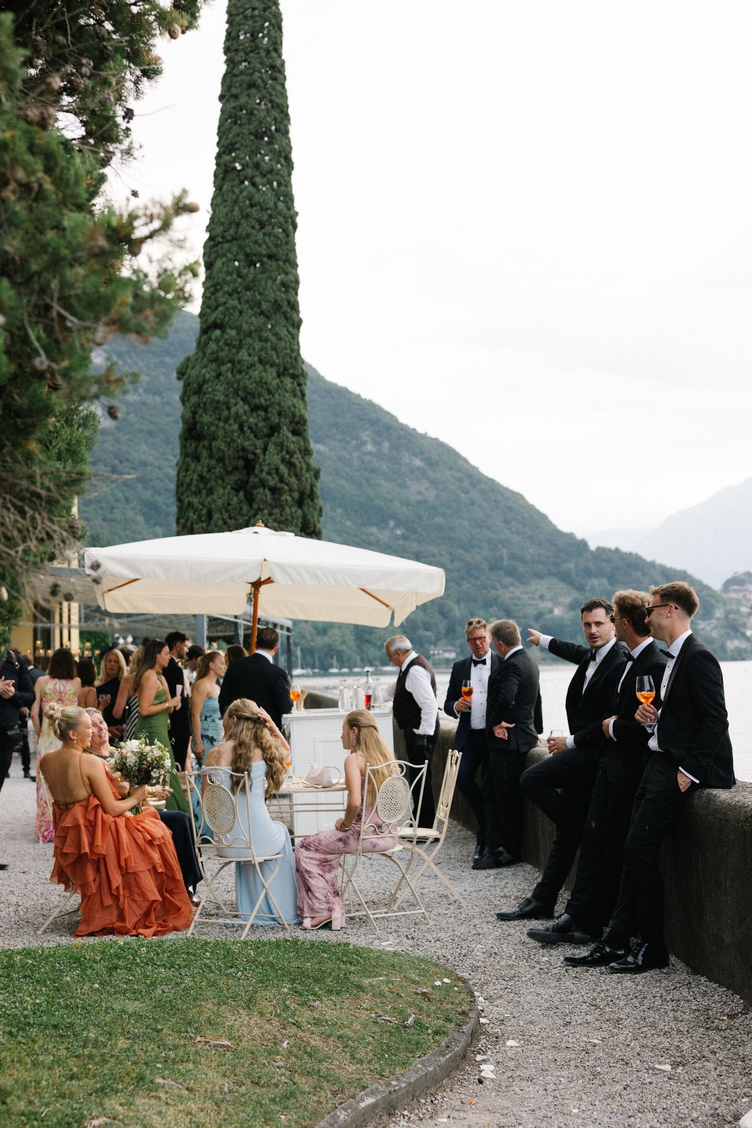 Elegant wedding cocktail hour by Lake Como, guests in formal attire mingling outdoors with mountain views.
