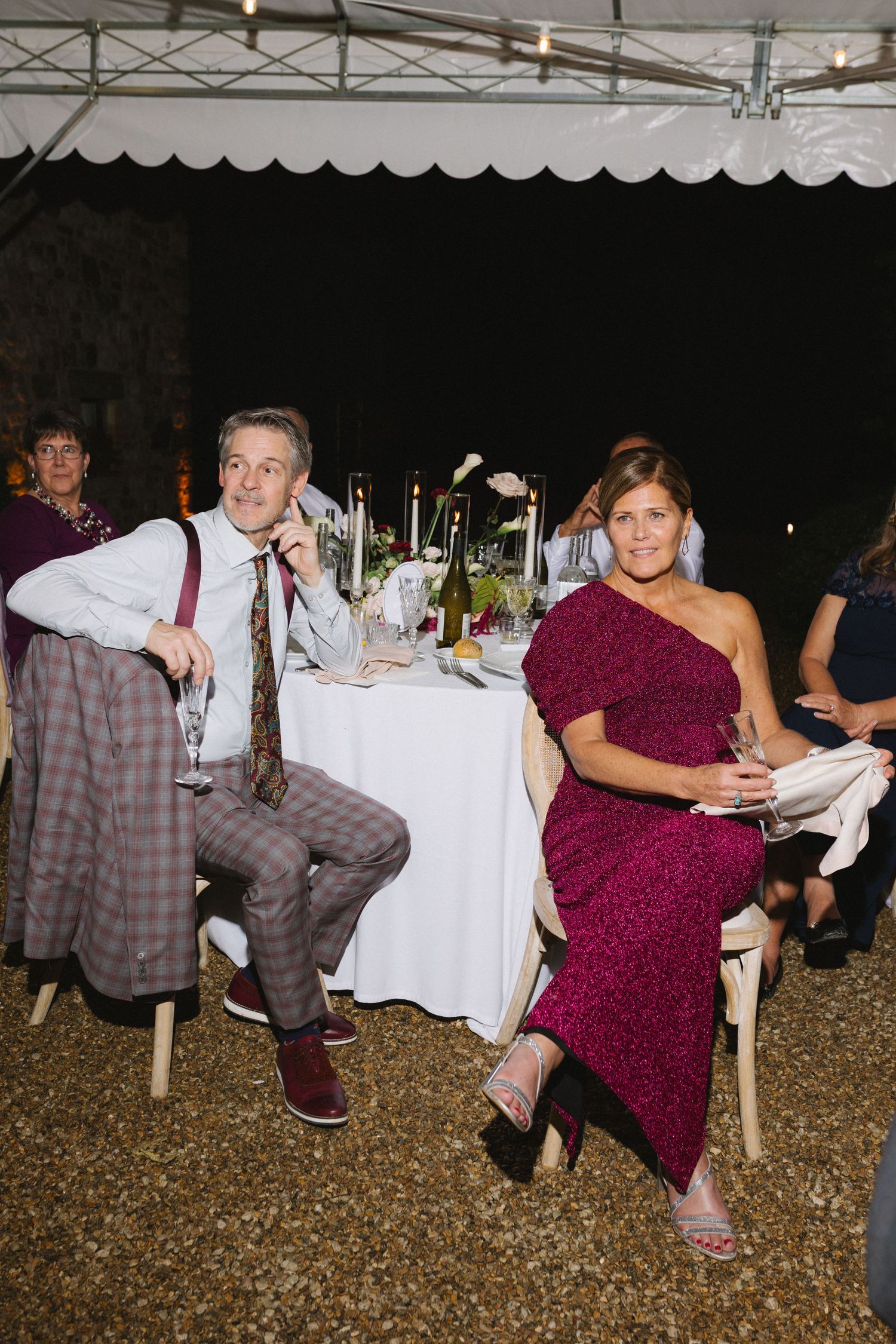 Guests in formal attire seated at an outdoor wedding reception table at night, woman in magenta dress, man in plaid suit.