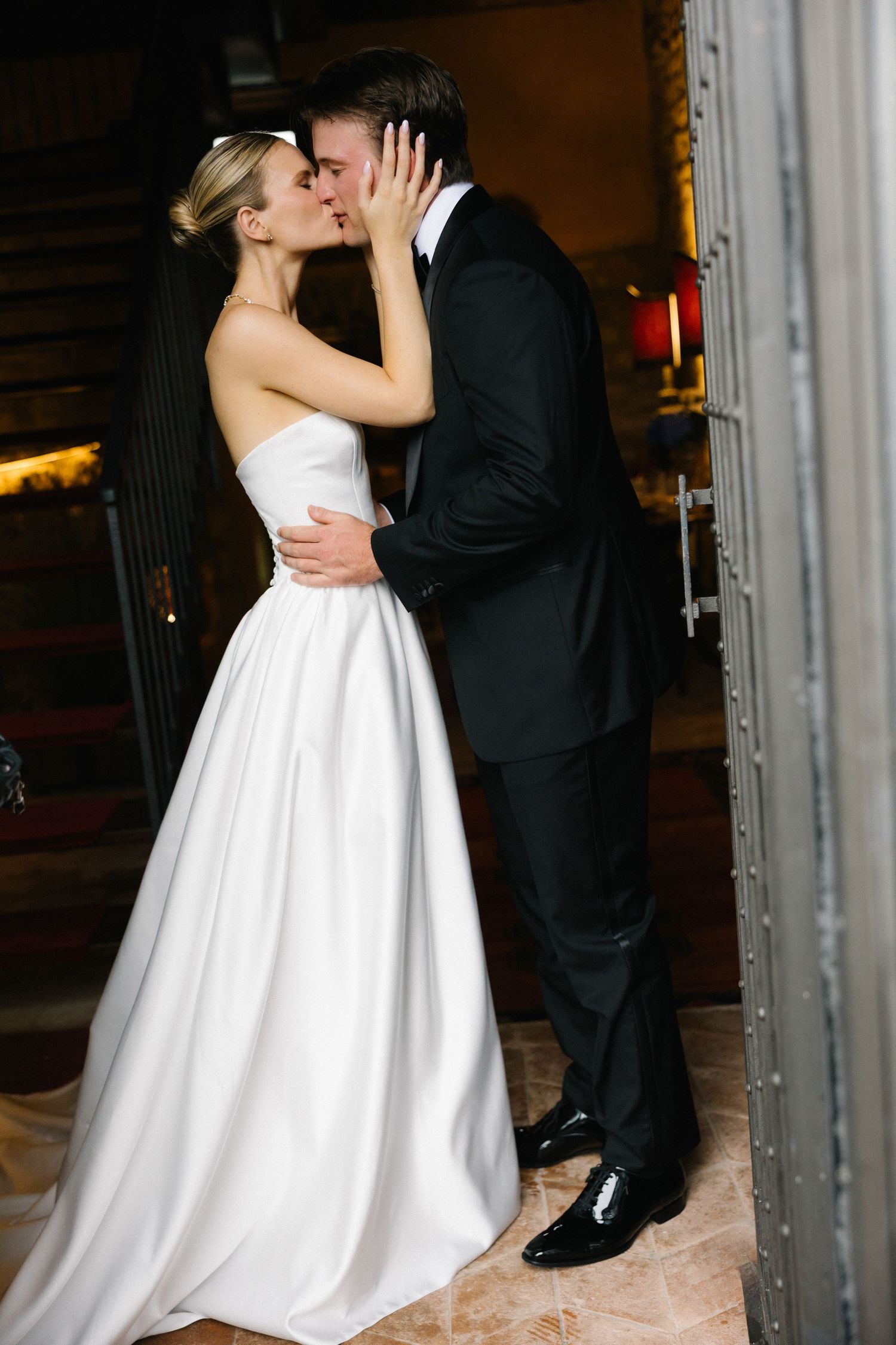 A bride in a white strapless gown and groom in black tuxedo share a romantic kiss at their wedding ceremony.
