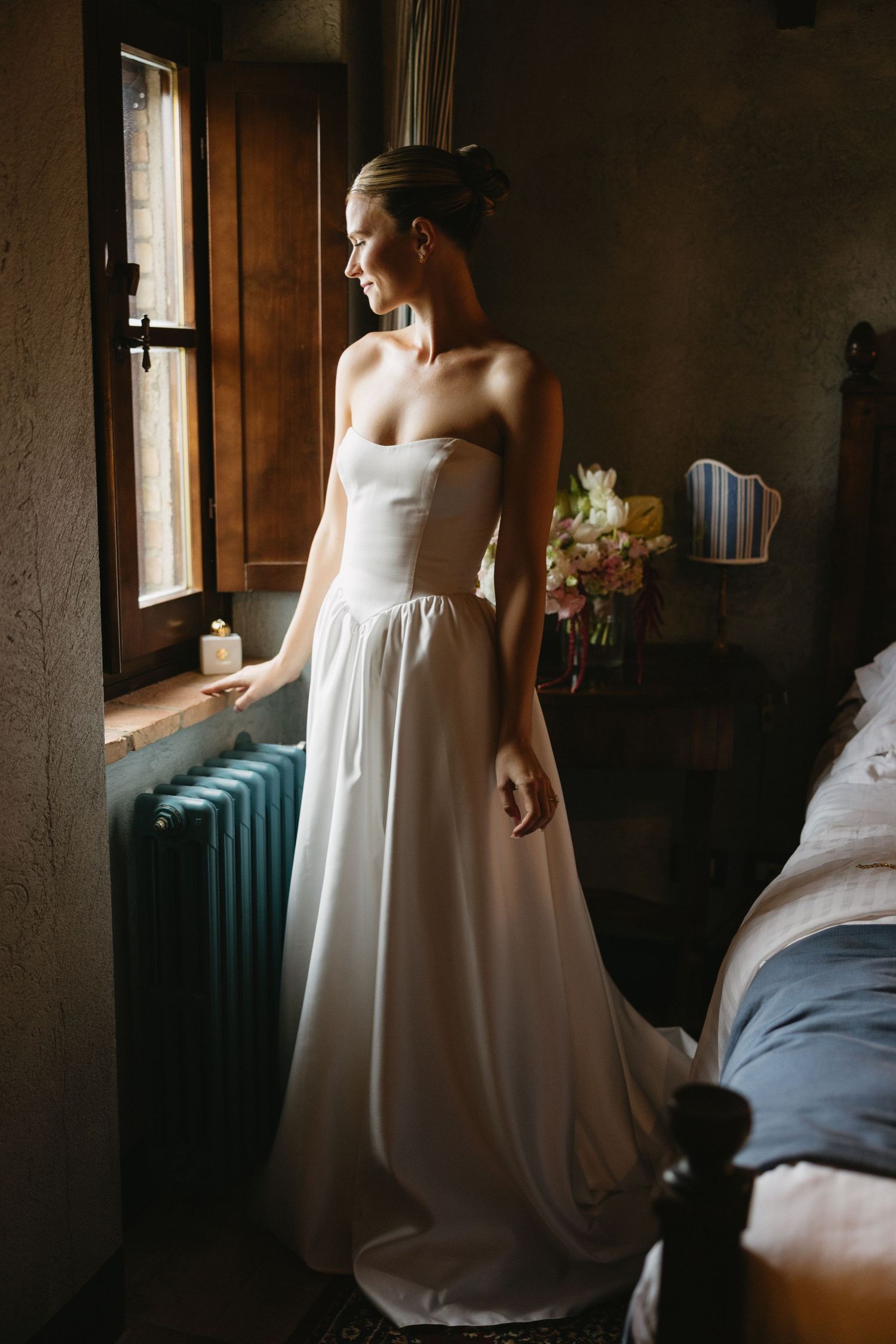 Bride in strapless white wedding gown standing by a rustic window in a dimly lit room with flowers in background.