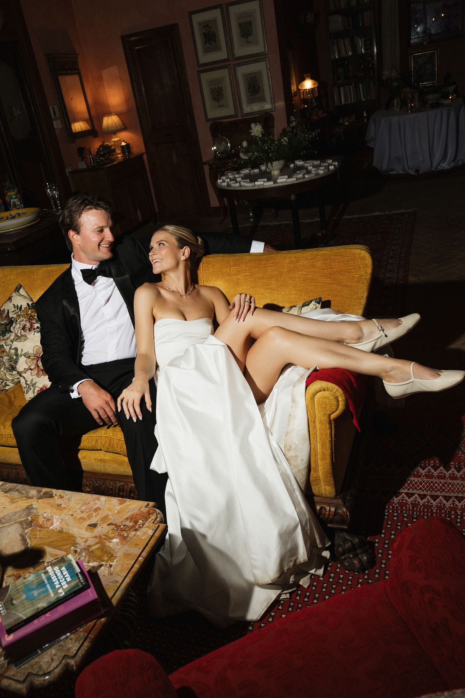 Bride and groom relaxing on a yellow sofa, she in a white gown with legs up, he in a black tuxedo, smiling at each other.
