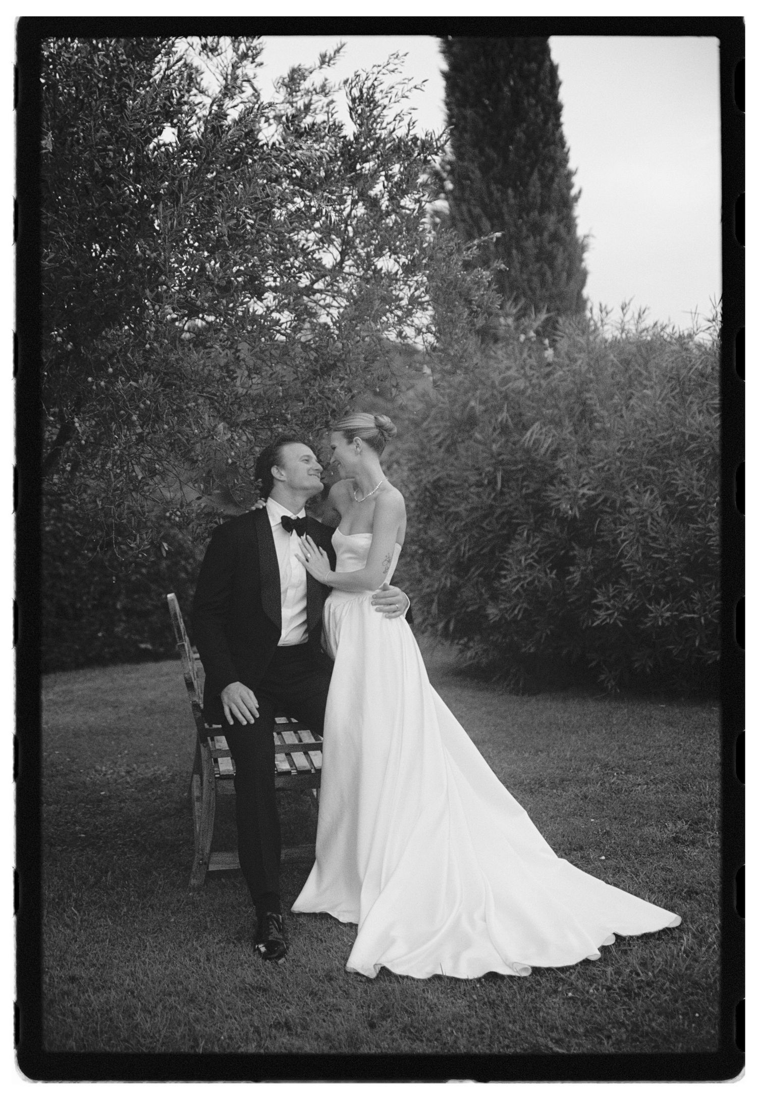 Black and white wedding photo of bride and groom embracing in a garden with cypress trees.