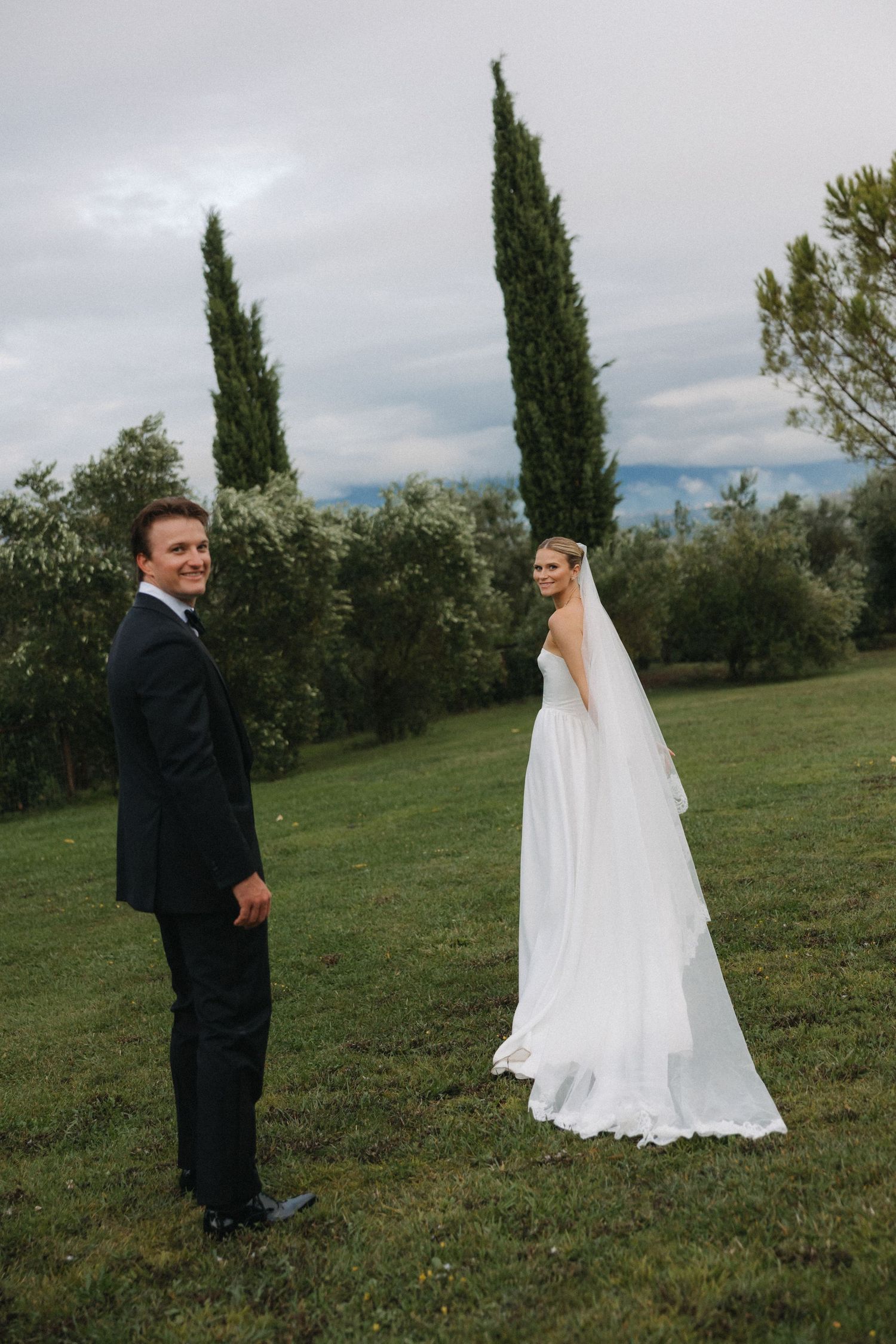 A bride in a white gown and groom in a dark suit pose on a green lawn with tall cypress trees in Tuscany.