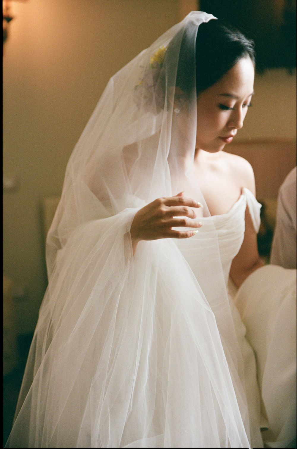 Elegant Asian bride in white off-shoulder gown and cathedral veil preparing for ceremony in warm golden light.