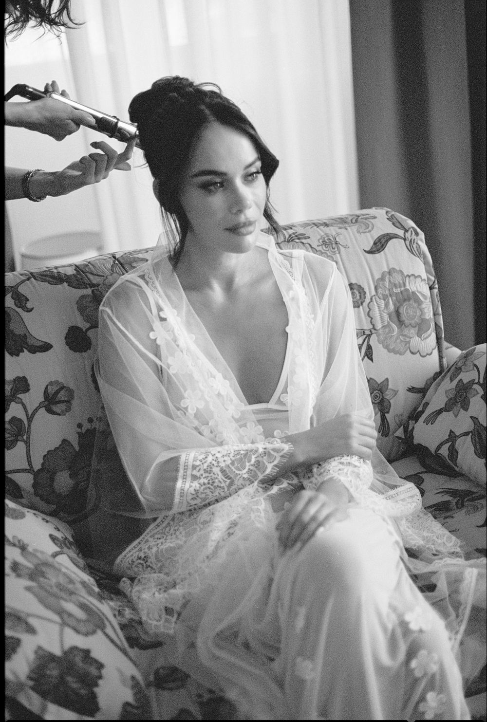 Bride in white lace robe getting hair styled before wedding ceremony.