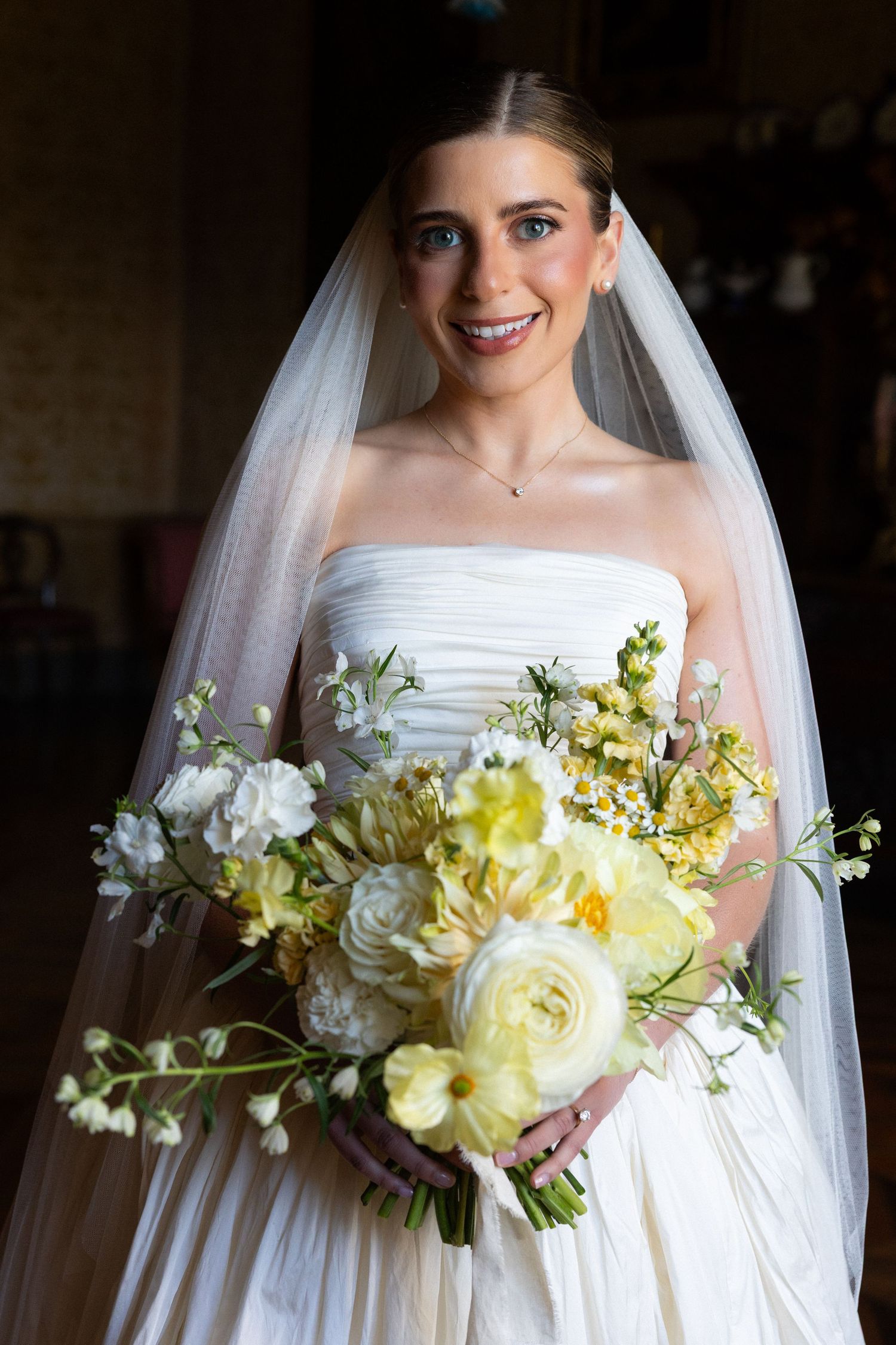 Smiling bride in strapless white gown and veil holding a lush bouquet of yellow and white flowers.