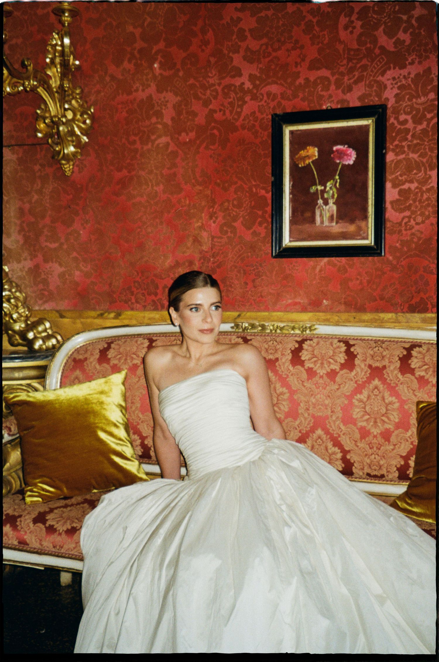 Elegant bride in strapless white ball gown seated on gold and red damask sofa in opulent baroque room.