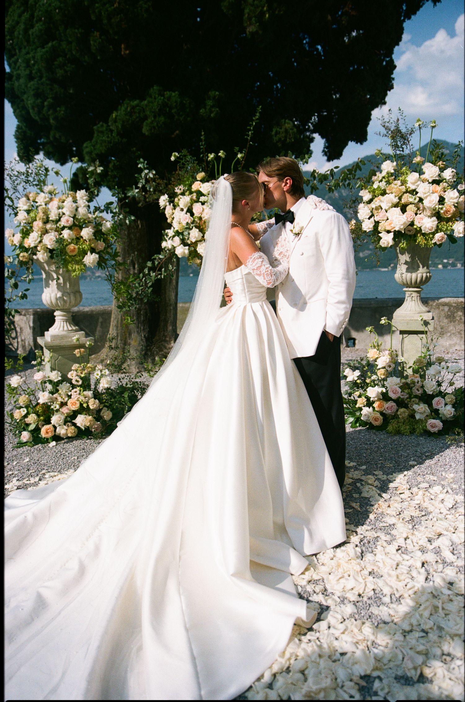 Bride and groom share a kiss at an outdoor wedding ceremony surrounded by elegant floral arrangements near a lakeside venue.