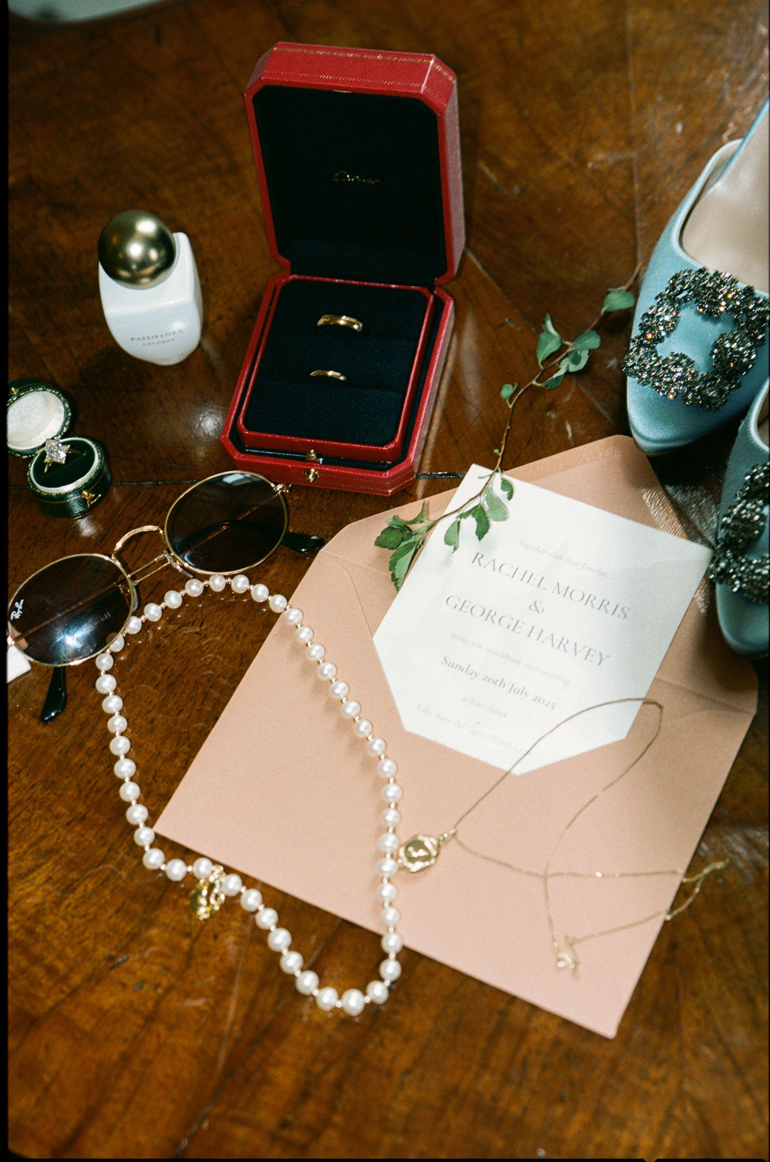 Wedding flat lay with Cartier ring box, pearl necklace, sunglasses, invitation, and jeweled heels on wooden table.