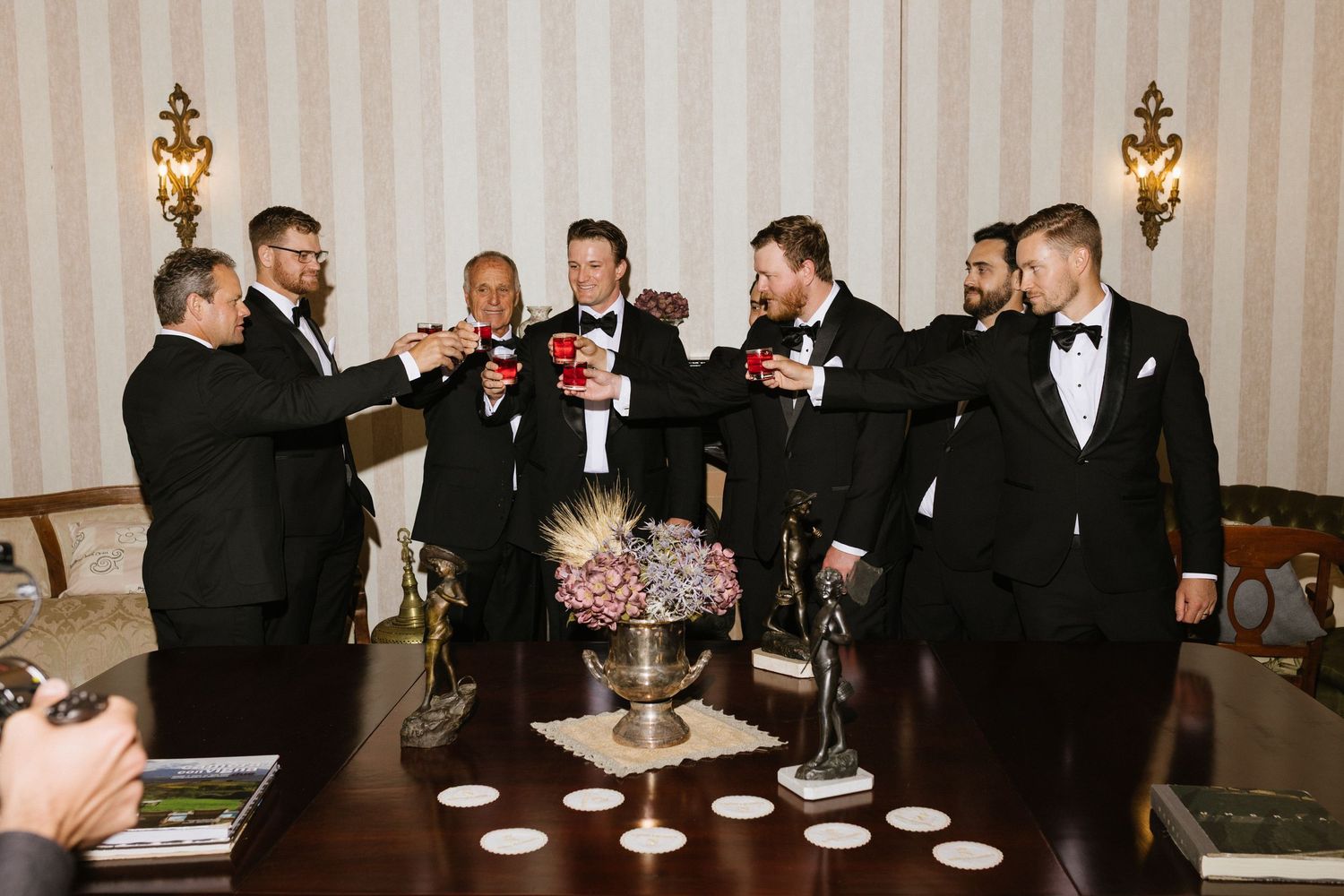 Groomsmen in black tuxedos raising glasses in a toast around a decorated table indoors.