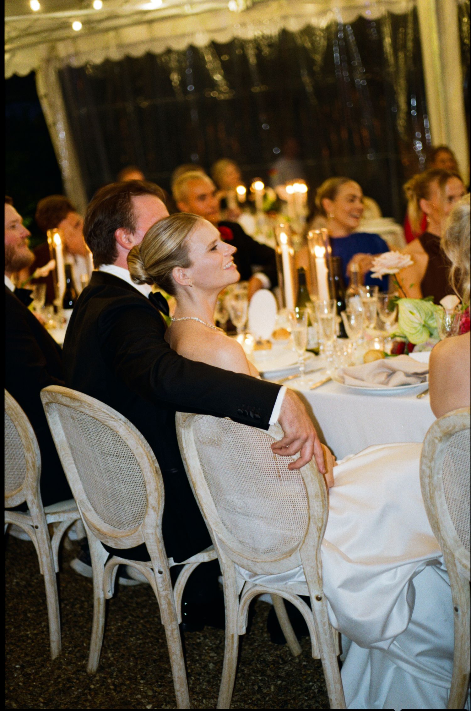 Elegant wedding reception dinner with bride and groom seated at candlelit table under white tent with guests.