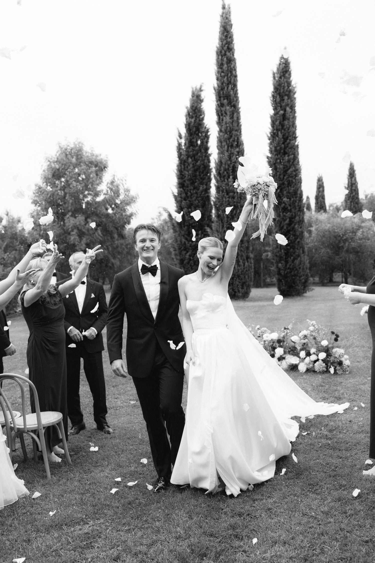 Black and white photo of a joyful bride and groom walking down the aisle as guests toss petals at outdoor wedding.