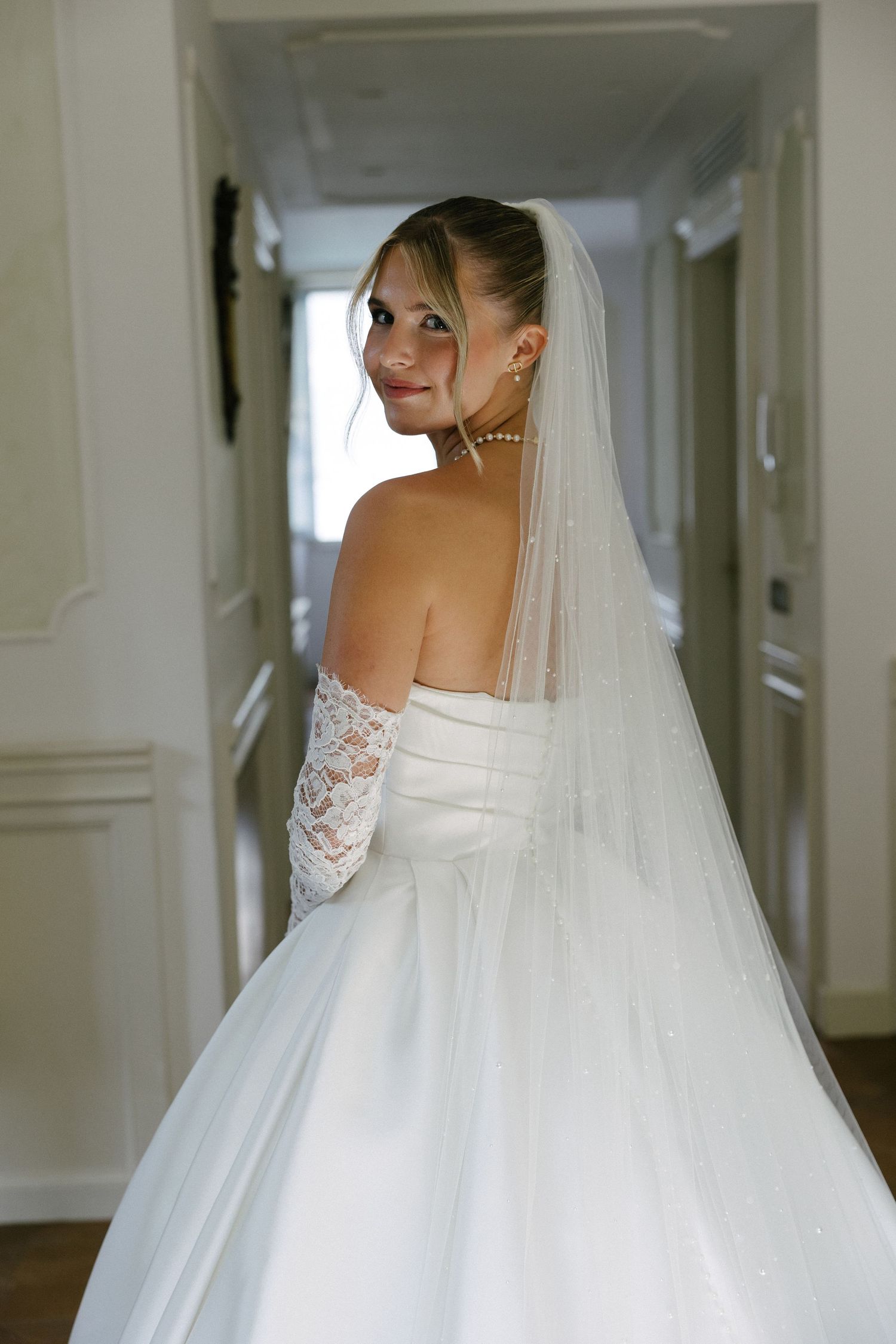 Bride in white strapless gown with lace gloves and veil smiles over shoulder in elegant hallway.