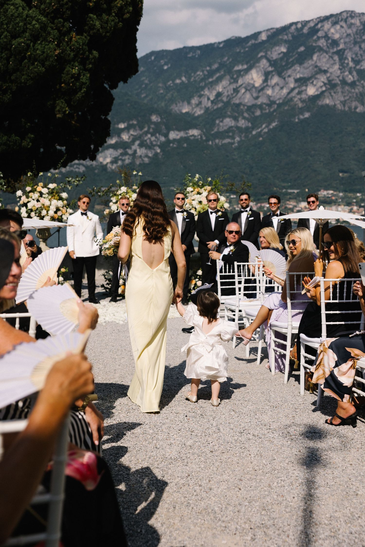 Flower girl walks down outdoor wedding aisle at Lake Como, Italy, with mountains and guests in background.
