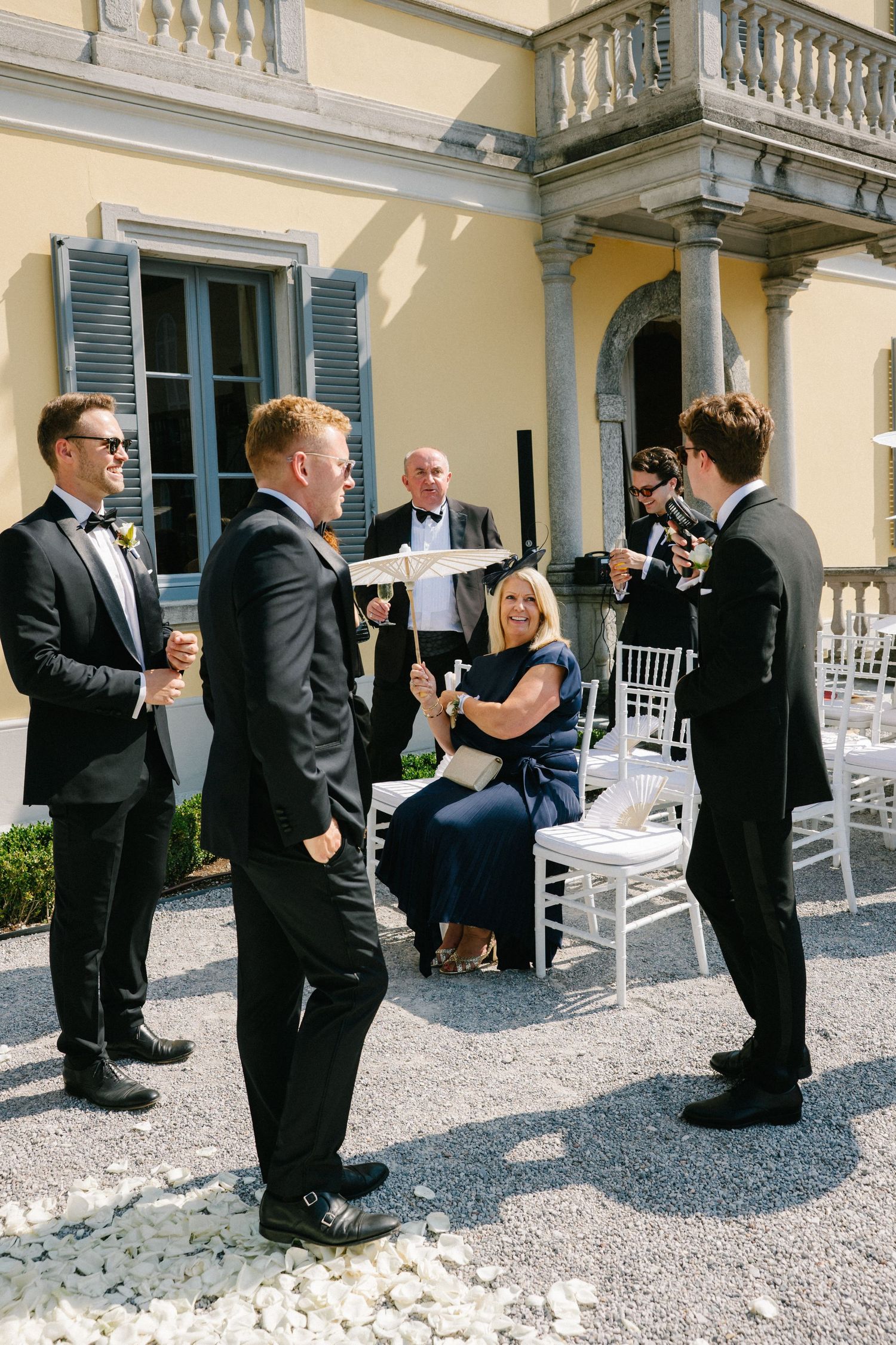 Groomsmen in black tuxedos chat with a seated woman in navy dress outside an elegant yellow villa wedding venue.