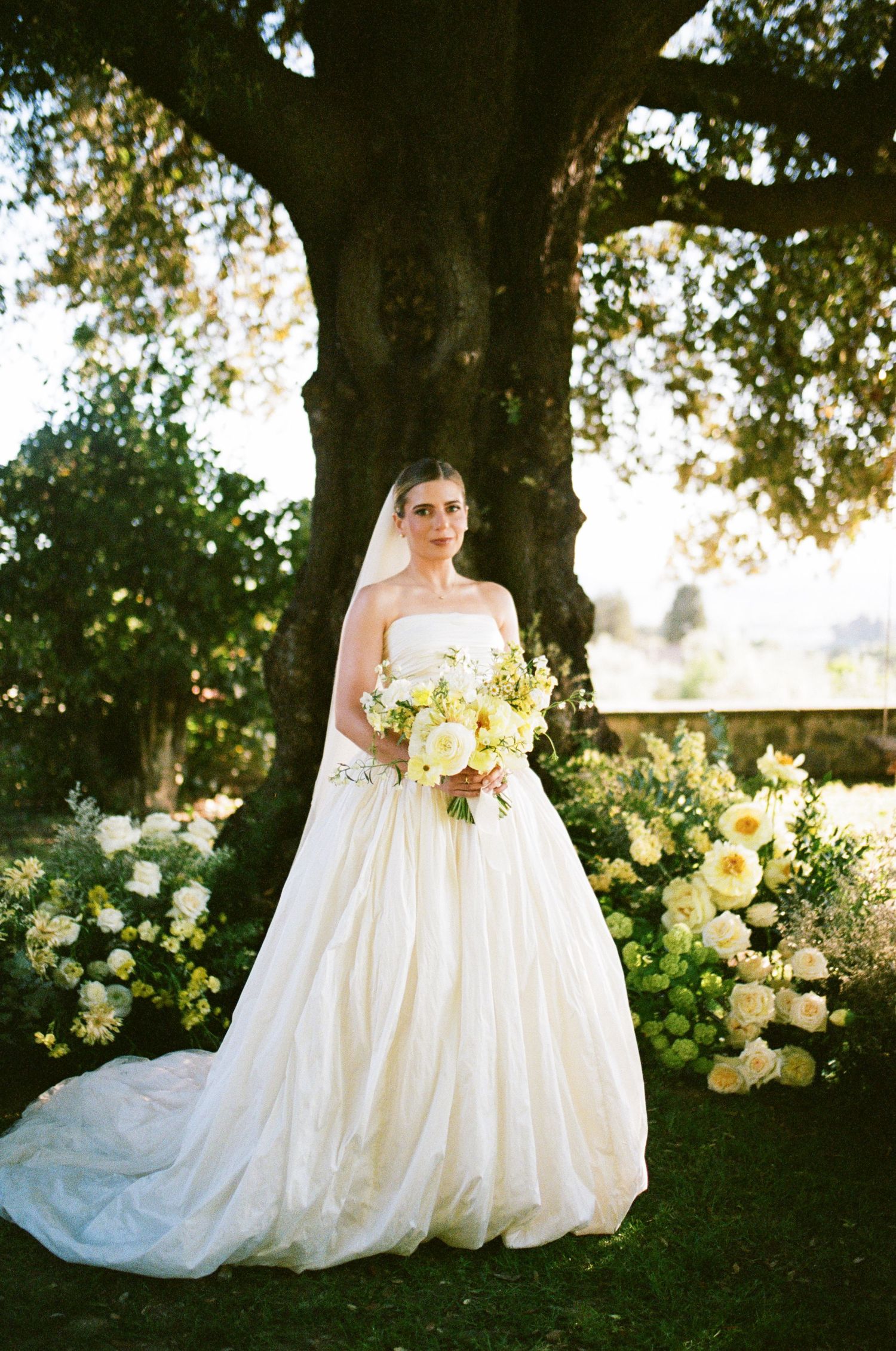 Bride in strapless ballgown holds white bouquet beneath a large tree surrounded by floral arrangements.